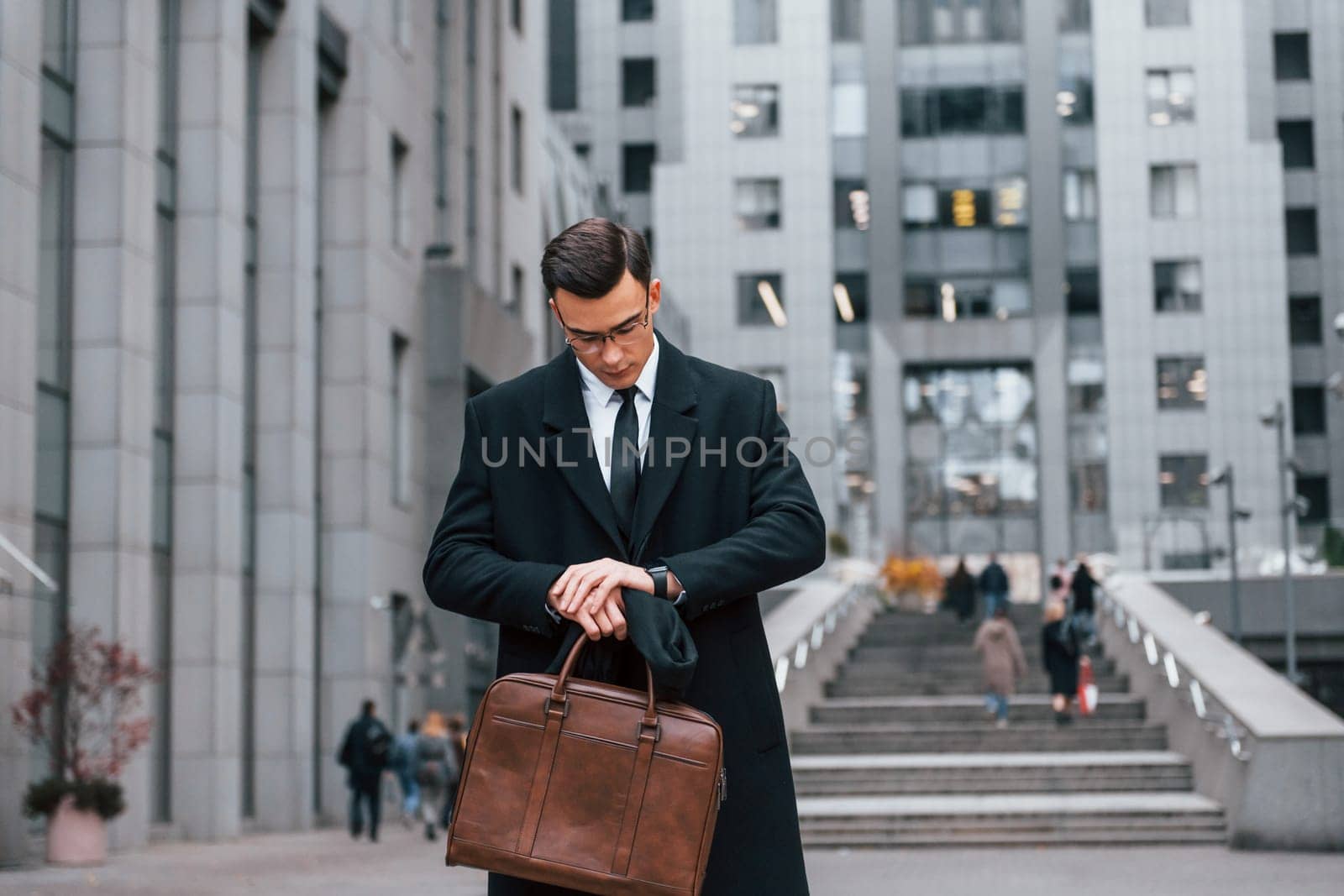 Looking at clock. Businessman in black suit and tie is outdoors in the city.