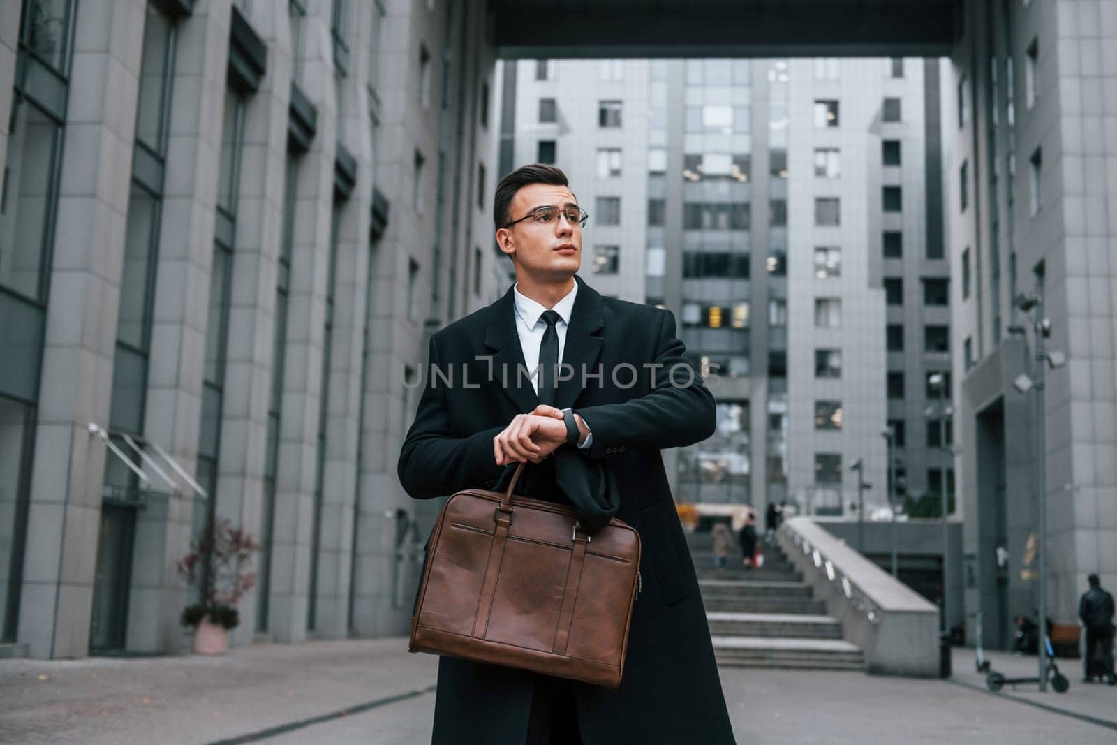Looking at clock. Businessman in black suit and tie is outdoors in the city.