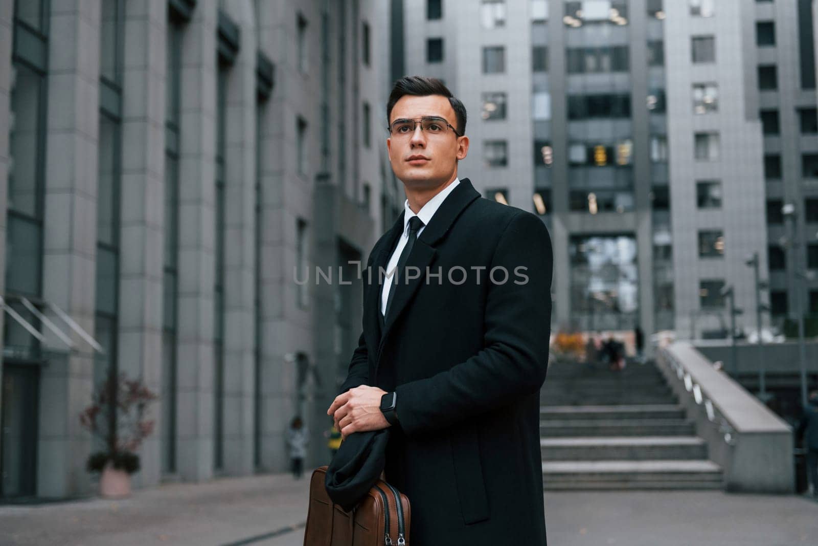 Front view. Businessman in black suit and tie is outdoors in the city.
