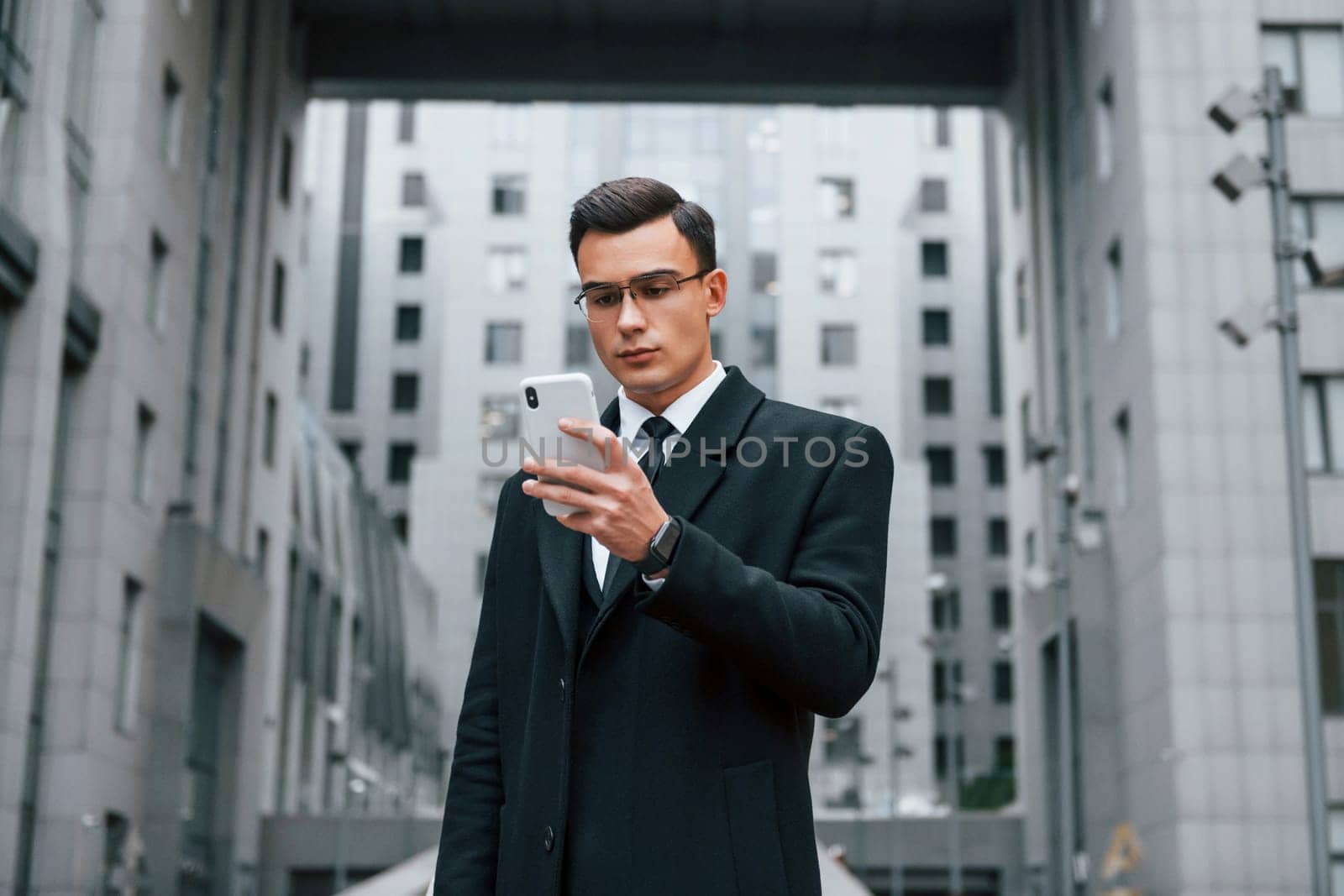 Phone in hand. Businessman in black suit and tie is outdoors in the city.