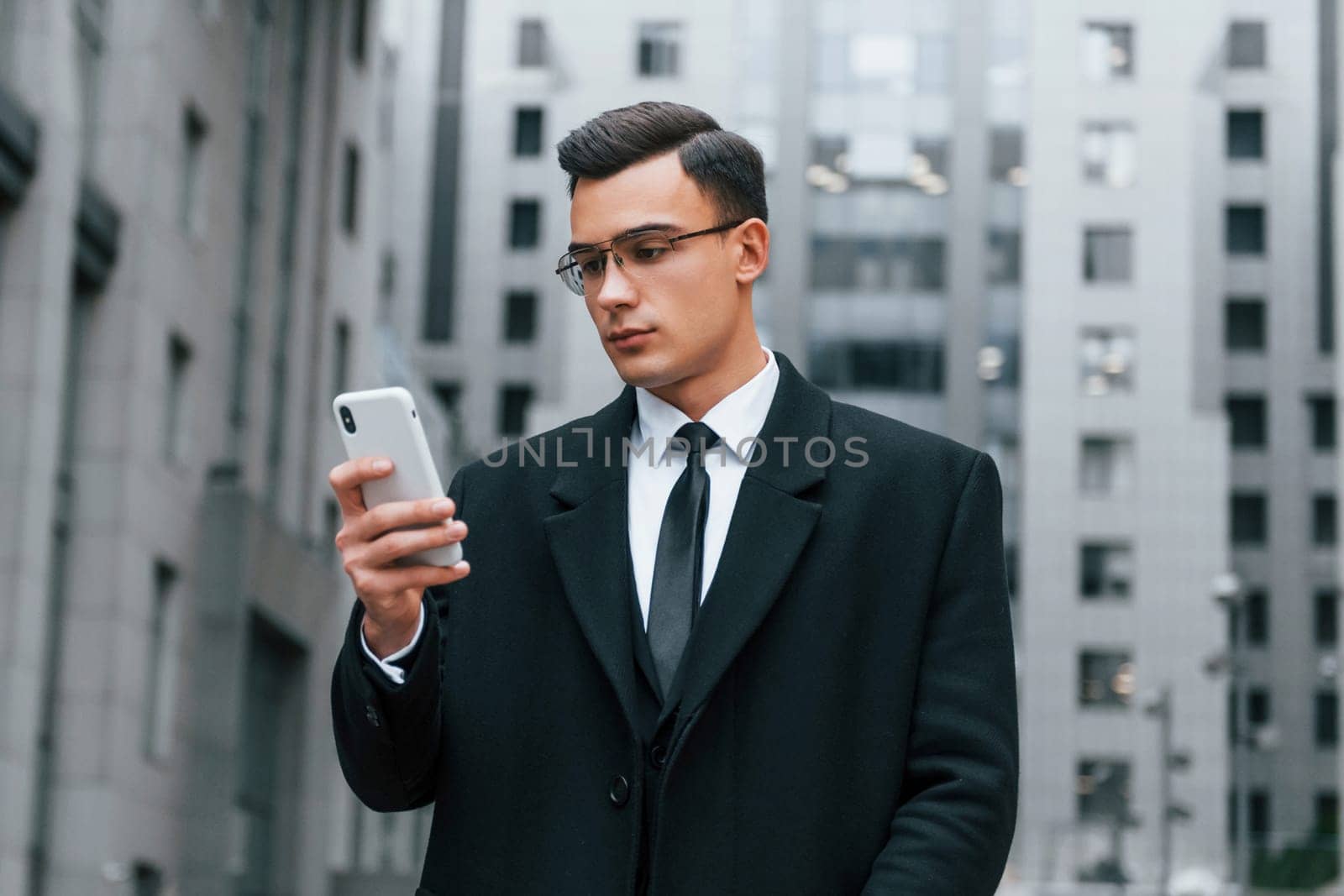 Phone in hand. Businessman in black suit and tie is outdoors in the city.