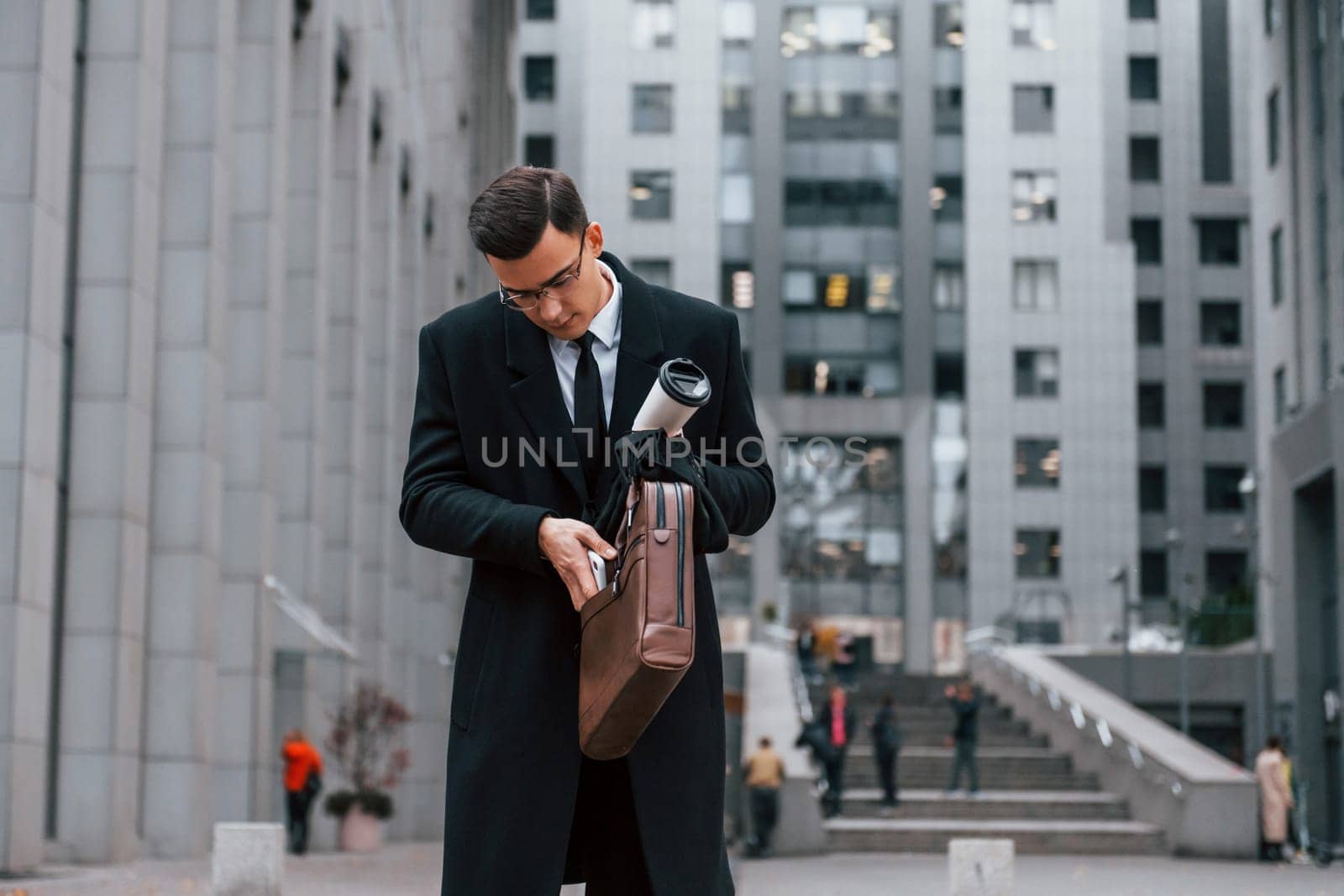 Putting phone in bag. Businessman in black suit and tie is outdoors in the city.