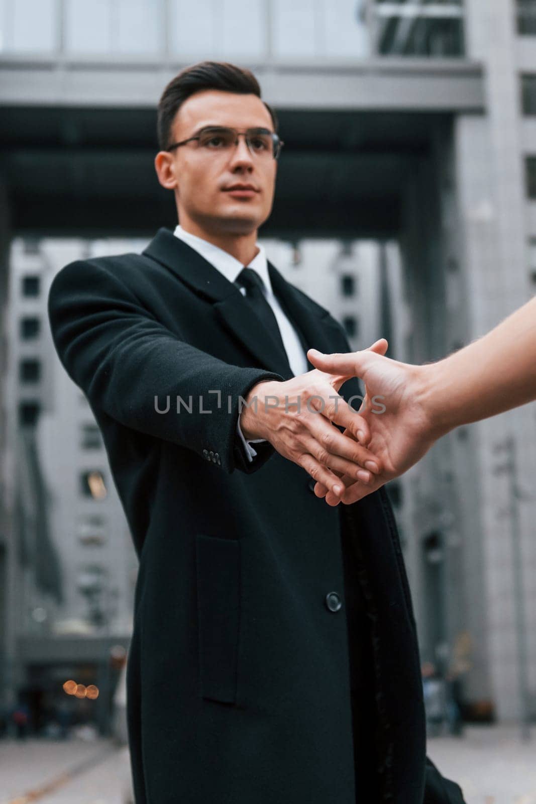 Doing handshake. Businessman in black suit and tie is outdoors in the city.