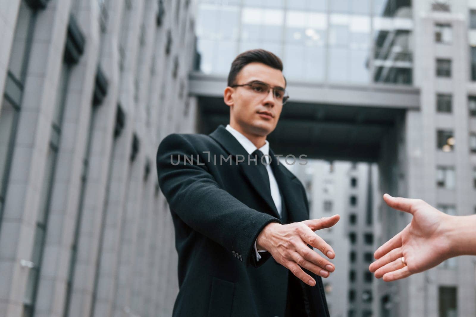 Doing handshake. Businessman in black suit and tie is outdoors in the city.