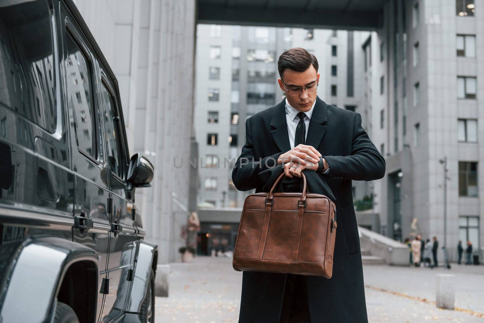 Standing near black car. Businessman in black suit and tie is outdoors in the city.