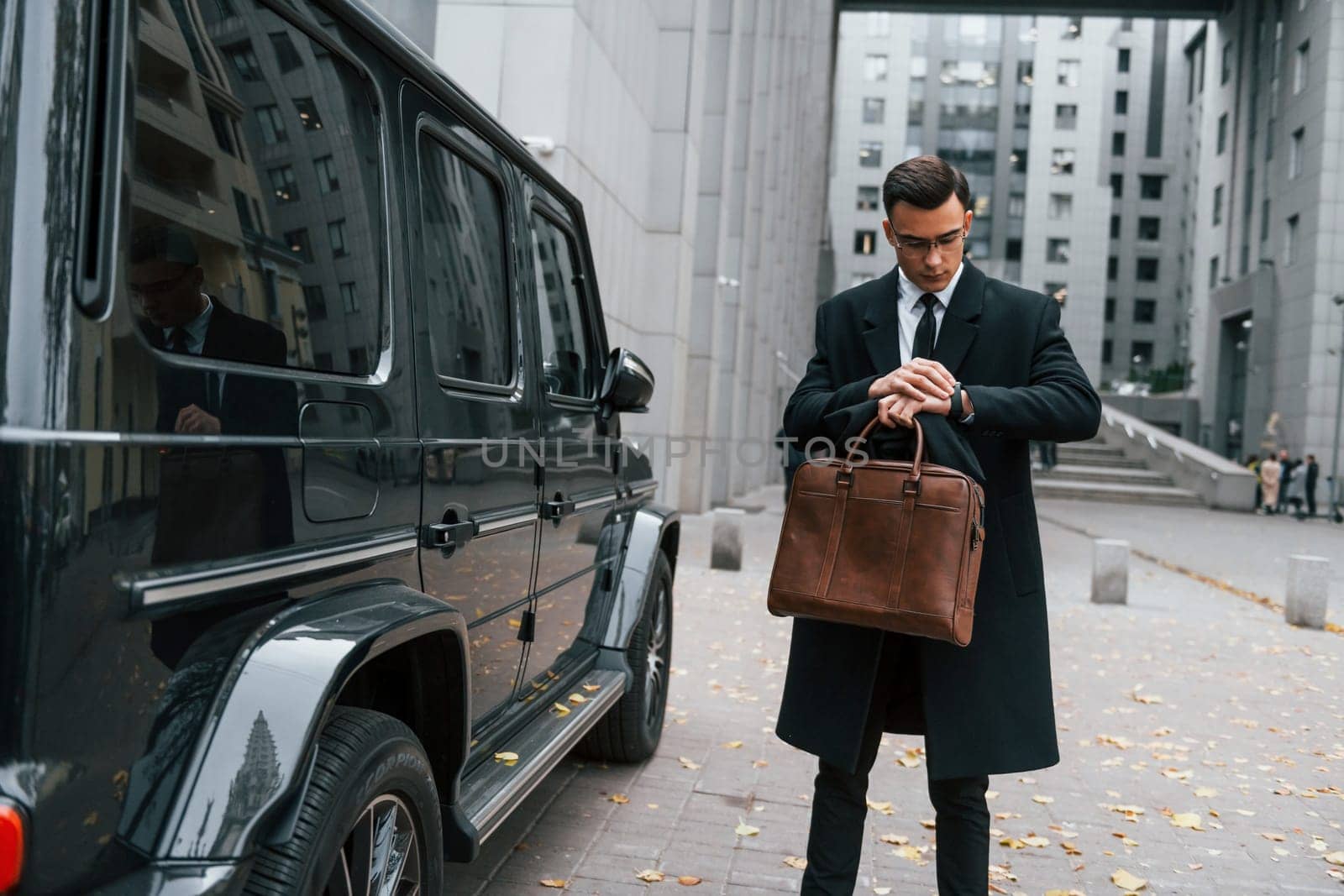 Standing near black car. Businessman in black suit and tie is outdoors in the city.