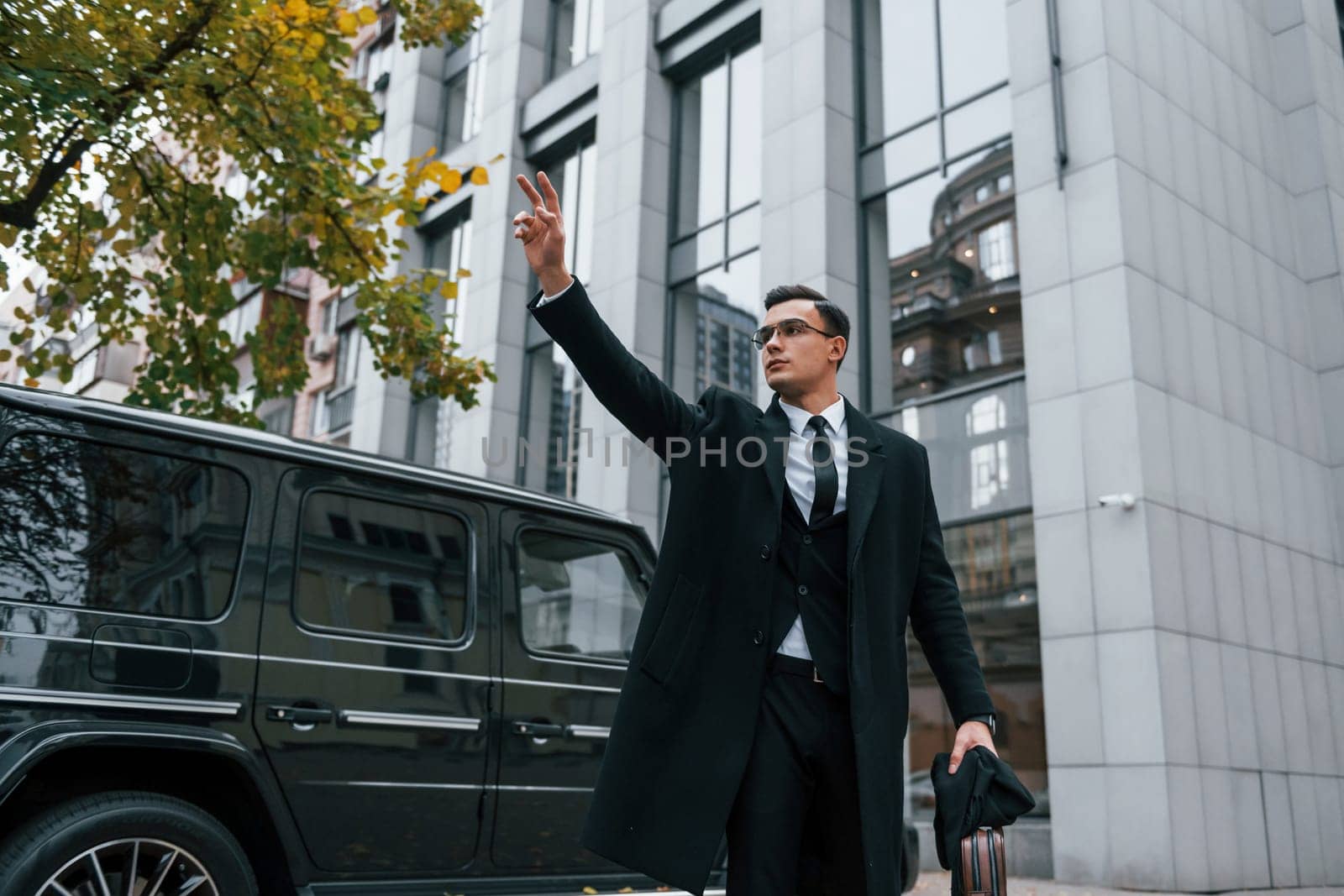 Standing against black car. Businessman in black suit and tie is outdoors in the city.