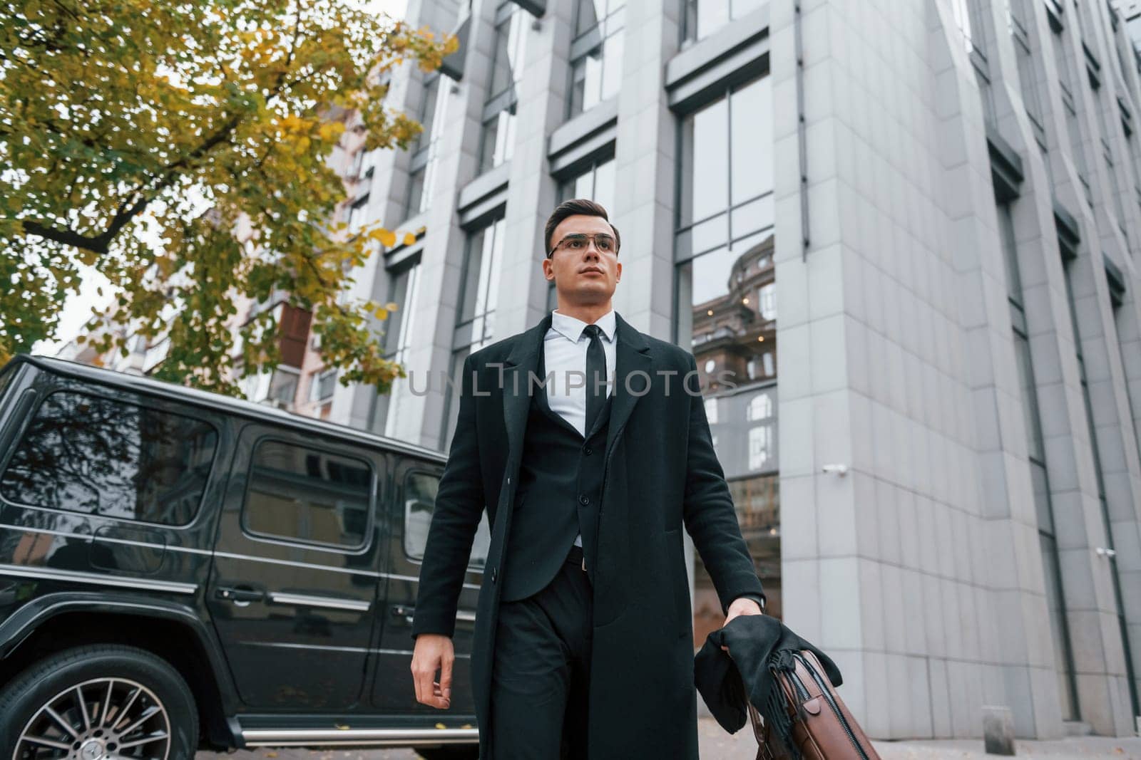 Standing against black car. Businessman in black suit and tie is outdoors in the city.