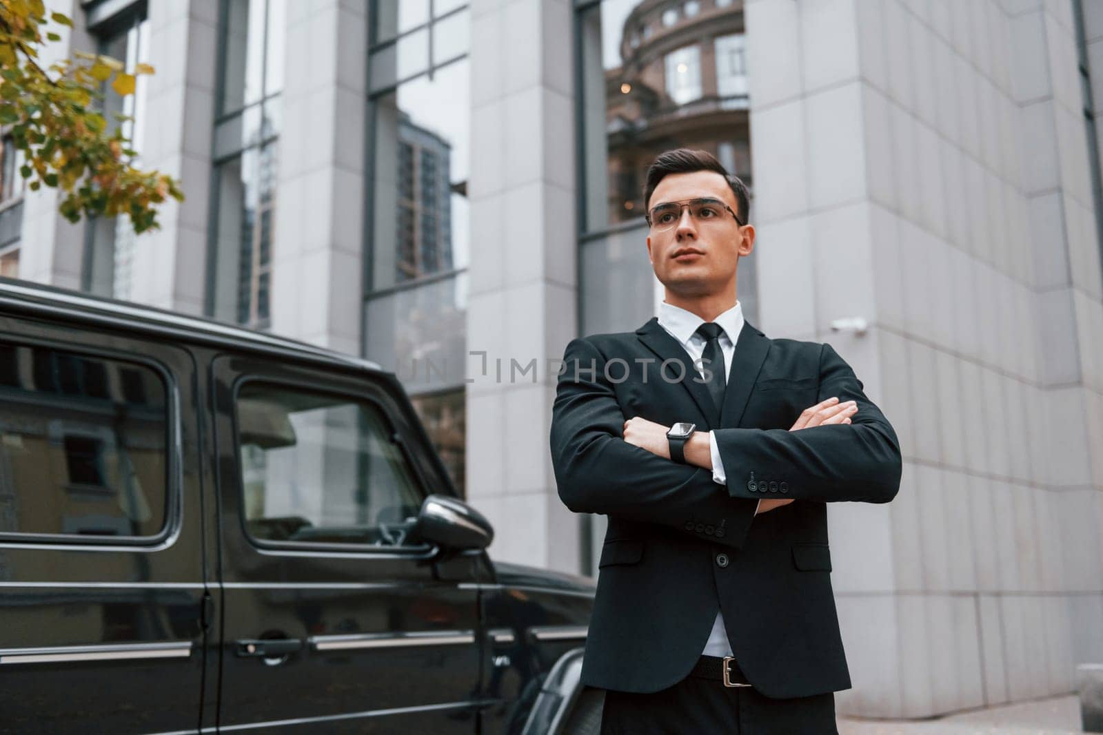 Standing with arms crossed. Businessman in black suit and tie is outdoors in the city by Standret