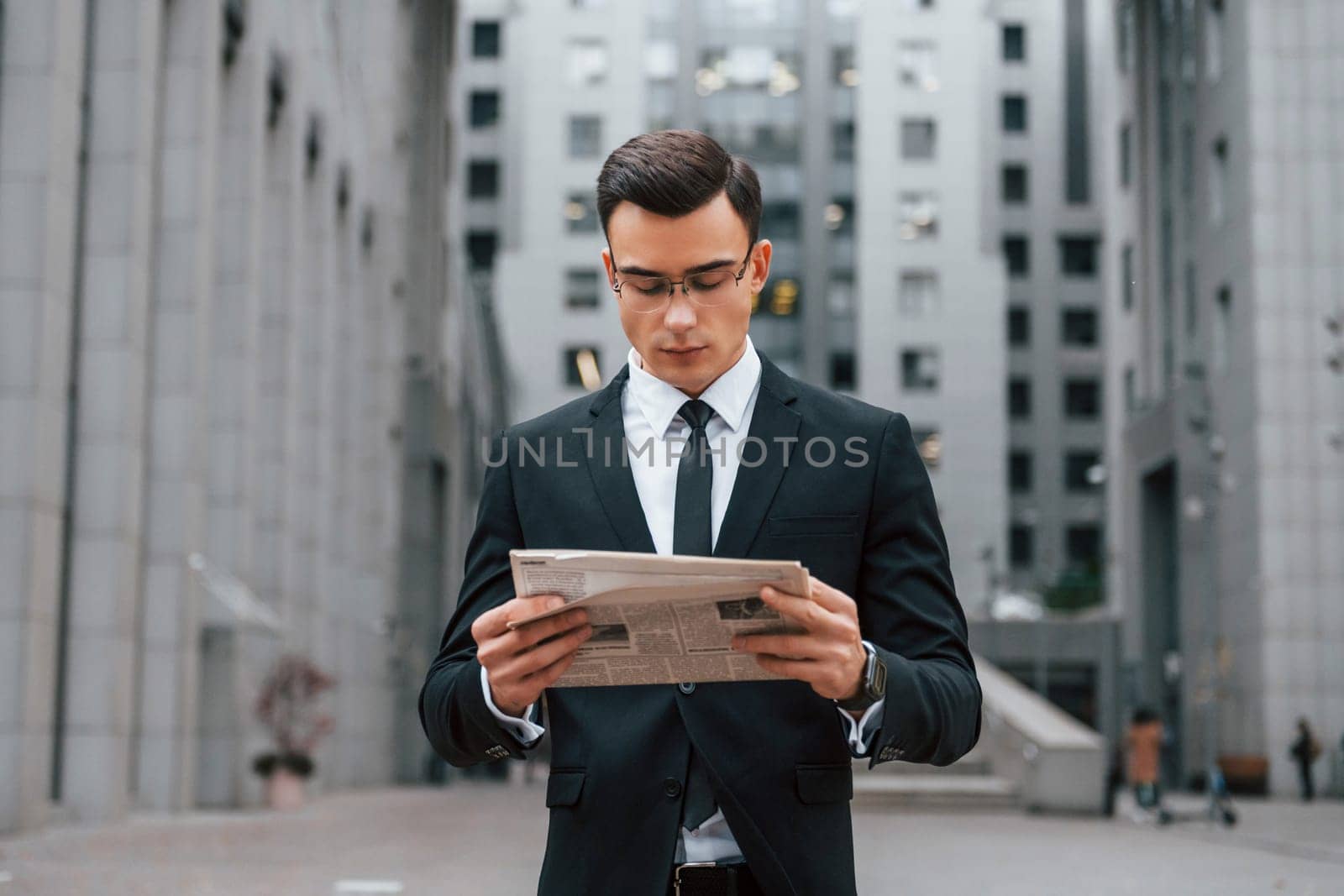 Reading some news. Businessman in black suit and tie is outdoors in the city.