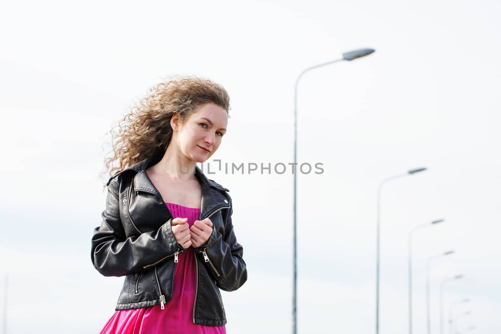 caucasian curly-haired woman in a black leather jacket and a pink dress against the background of a cloudy sky and pillars with lanterns