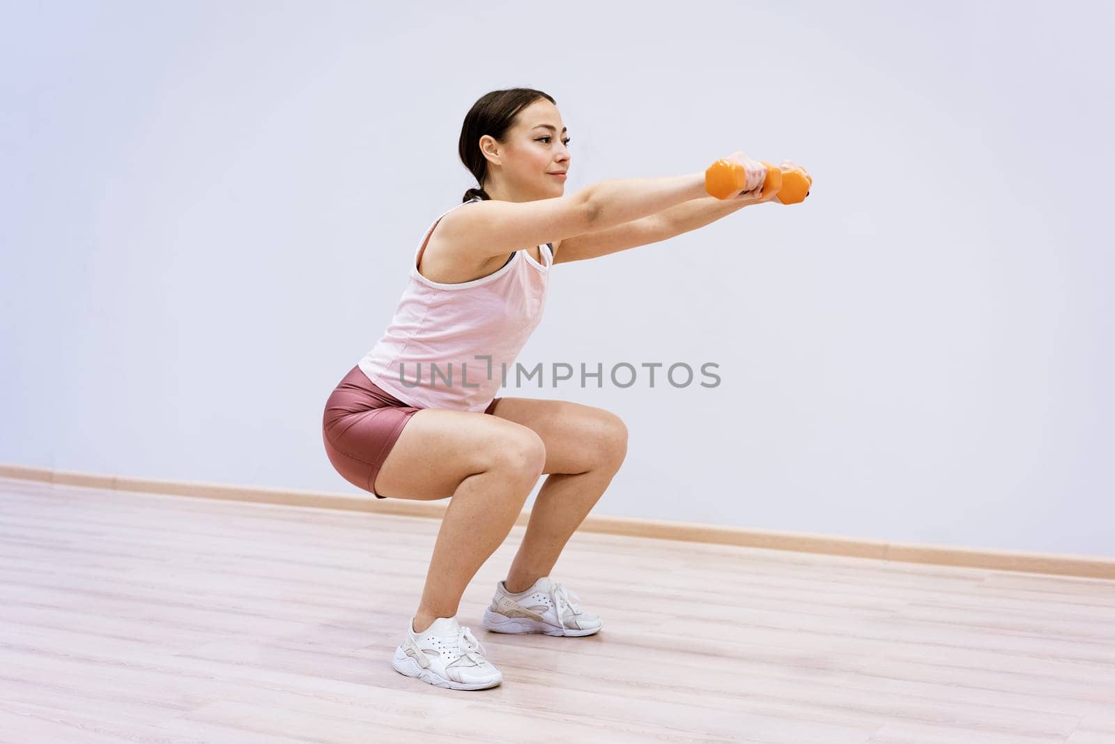 Woman squats with dumbbells in her hand against the background of a light wall by EkaterinaPereslavtseva