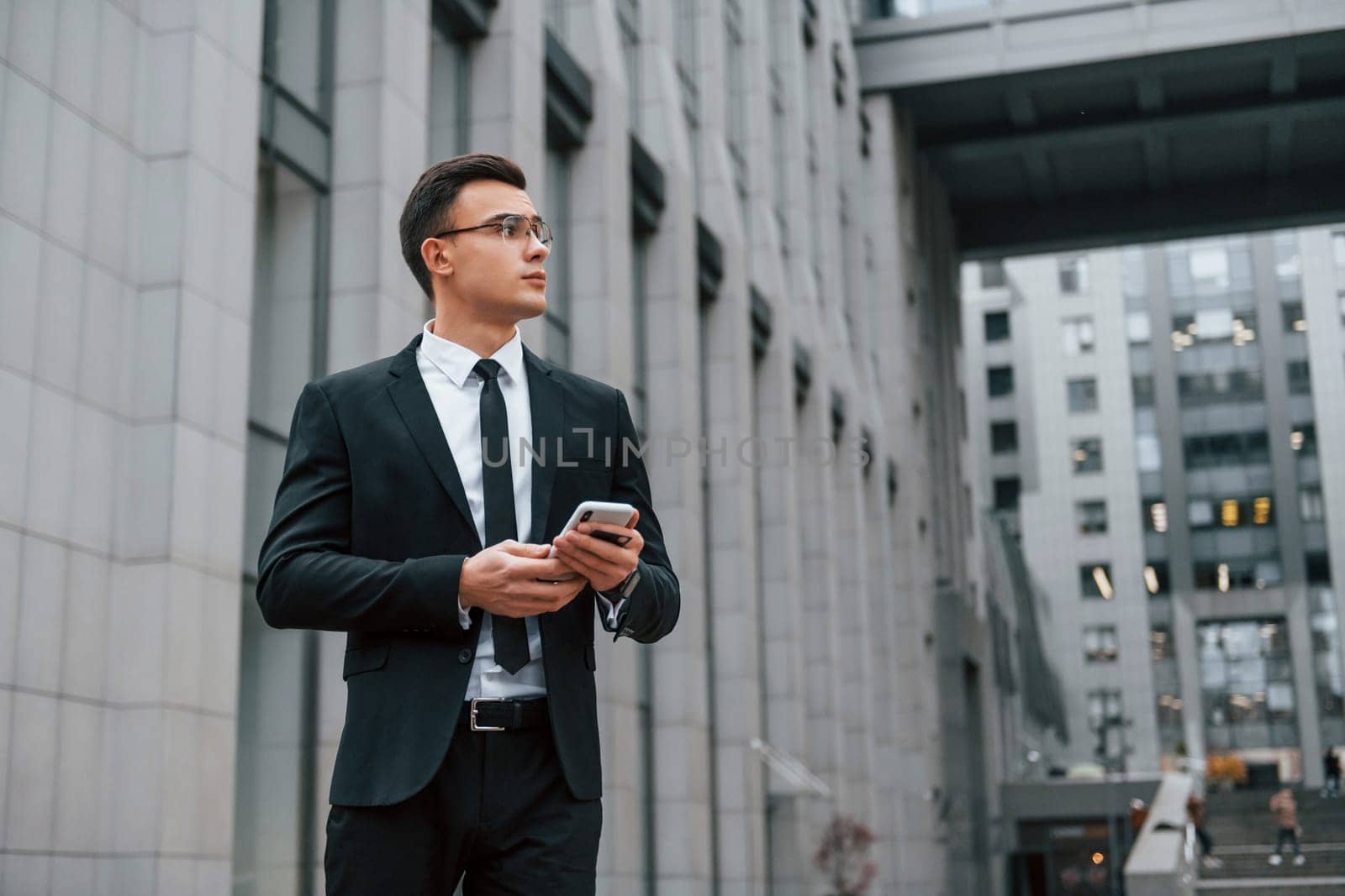 Standing and waiting. Businessman in black suit and tie is outdoors in the city.
