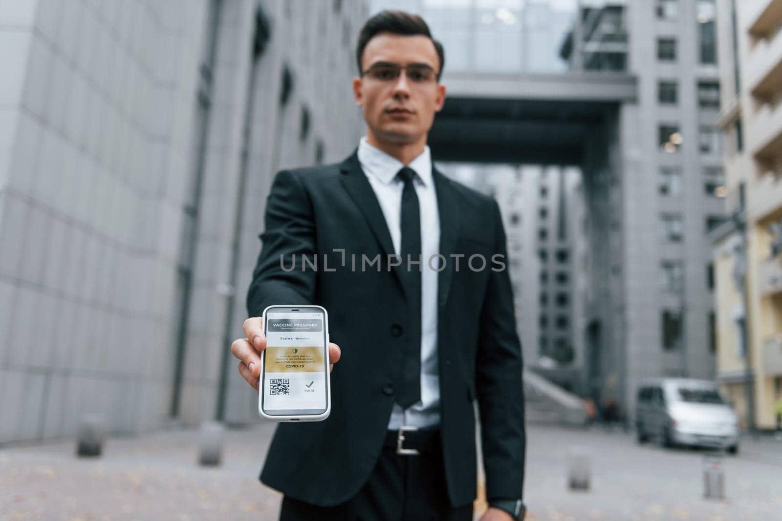 Holding phone with vaccination certificate. Businessman in black suit and tie is outdoors in the city.