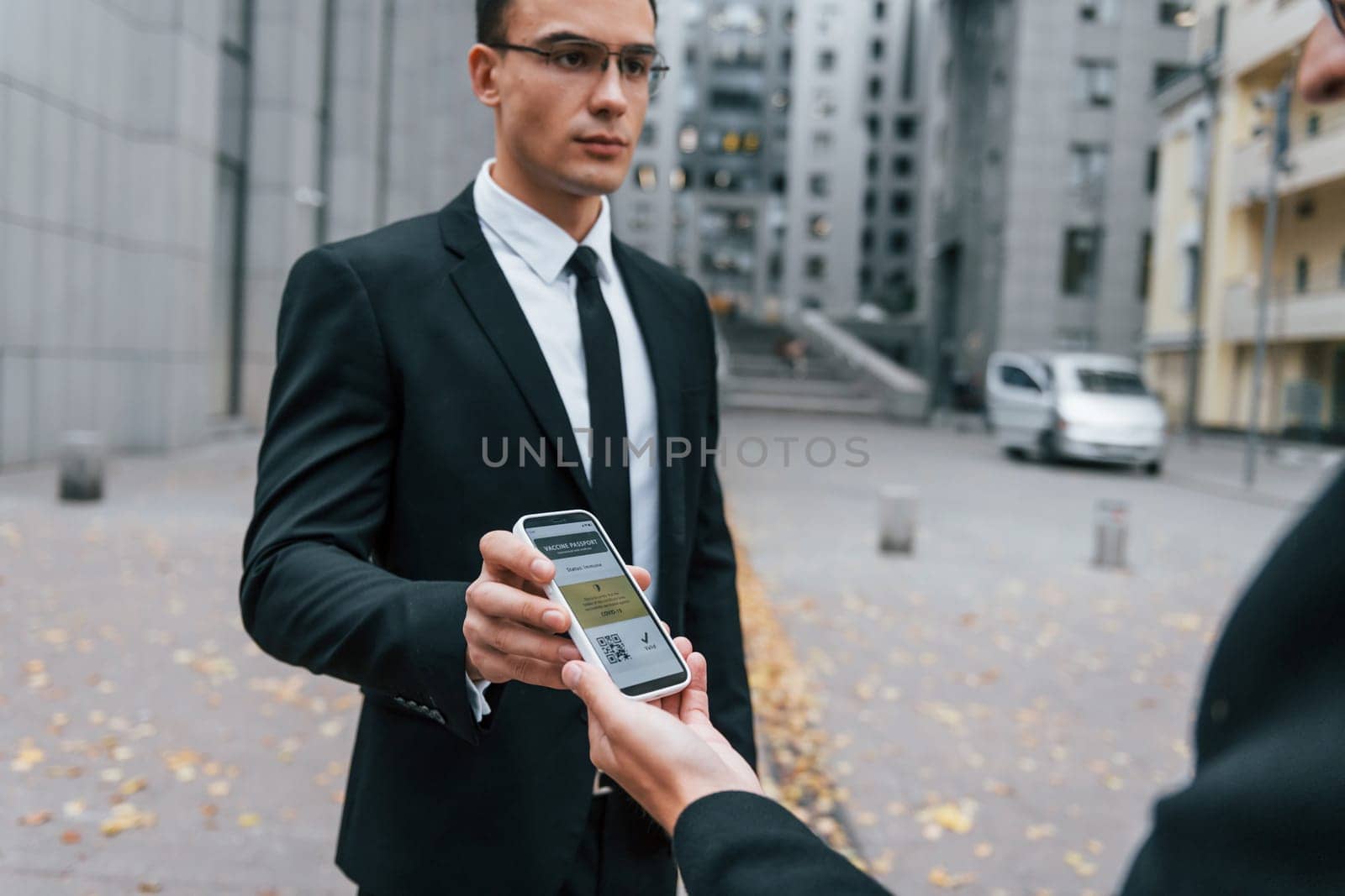 Holding phone with vaccination certificate. Businessman in black suit and tie is outdoors in the city.