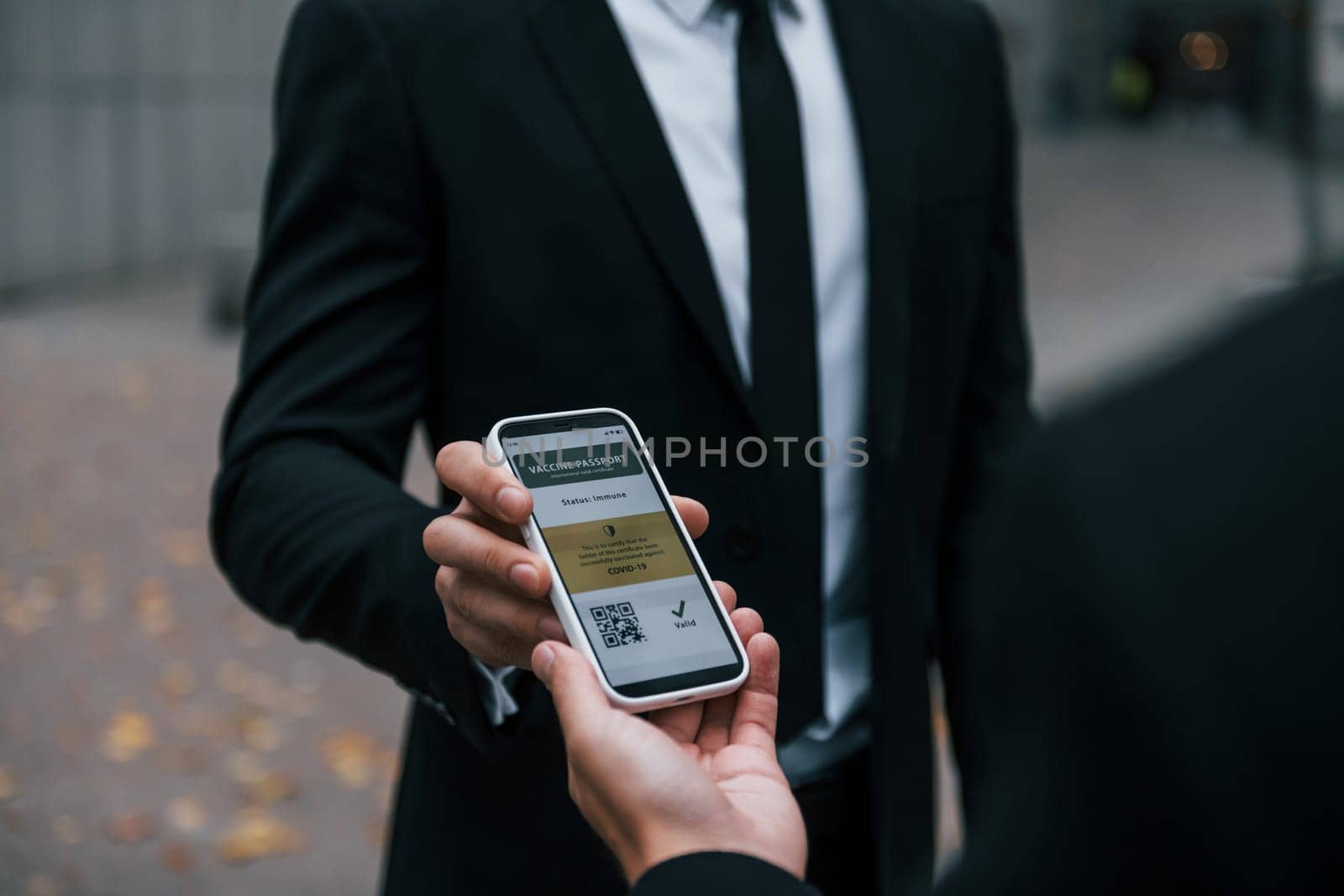 Holding phone with vaccination certificate. Businessman in black suit and tie is outdoors in the city.