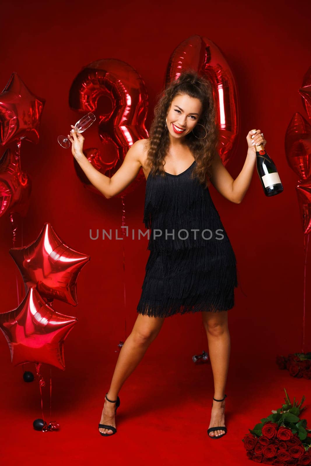 Happy woman with bottle of champagne on red background with red balloons. Cheerful mood at the holiday, anniversary of 30 years. A girl with long hair and bright red lips smiles with snow-white teeth