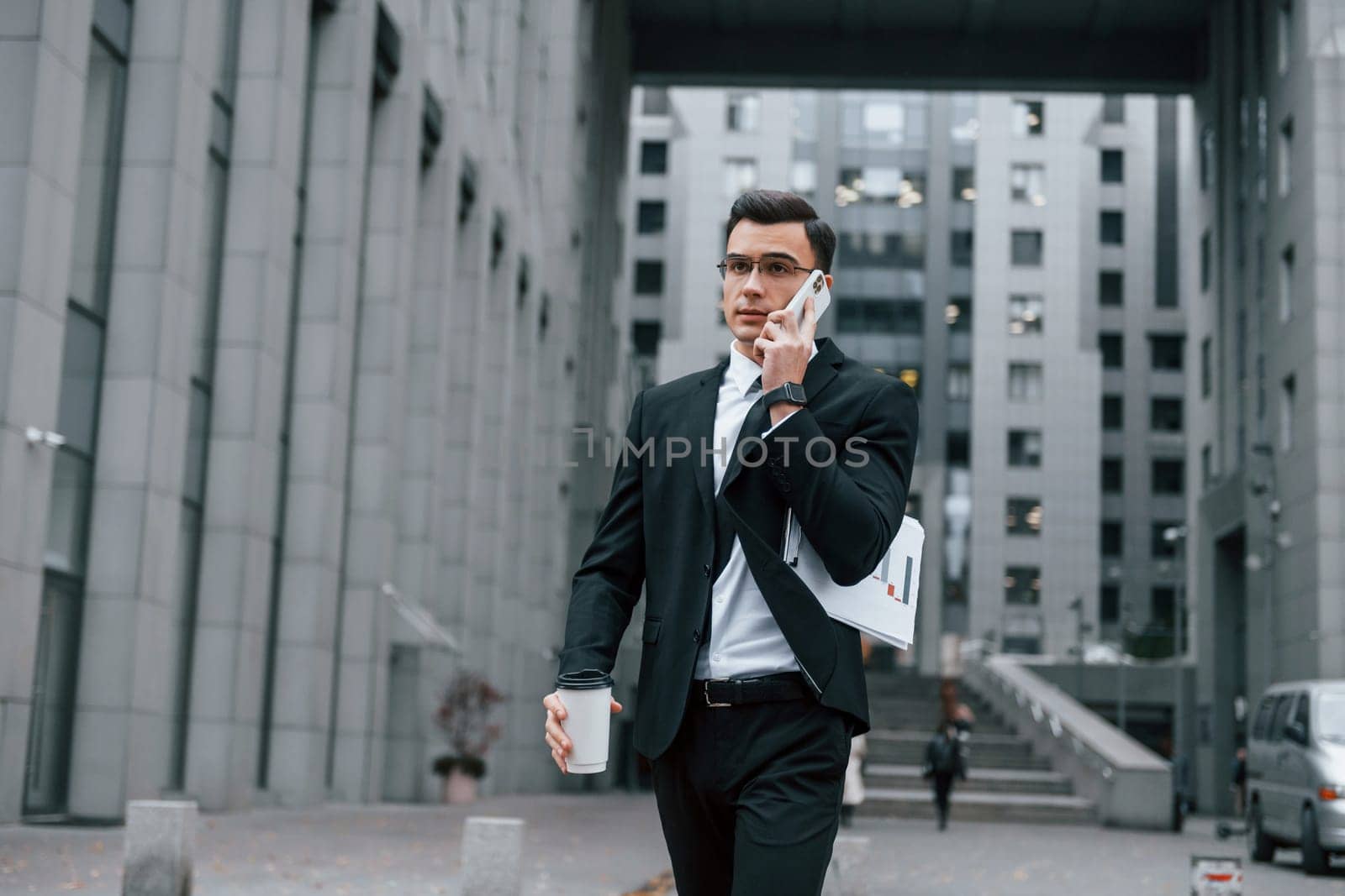 Walking and talking by phone. Businessman in black suit and tie is outdoors in the city.