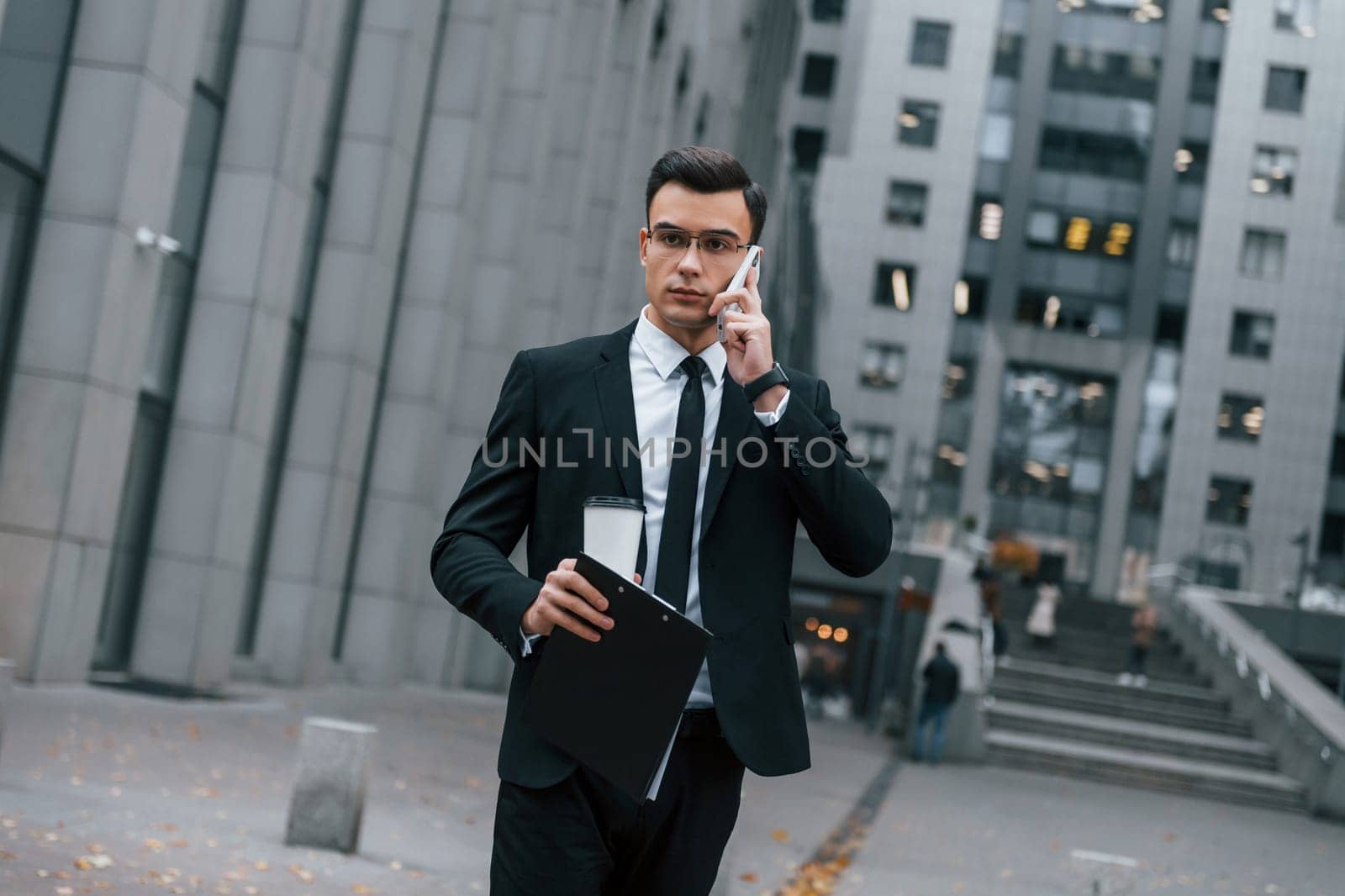 Walking and talking by phone. Businessman in black suit and tie is outdoors in the city.