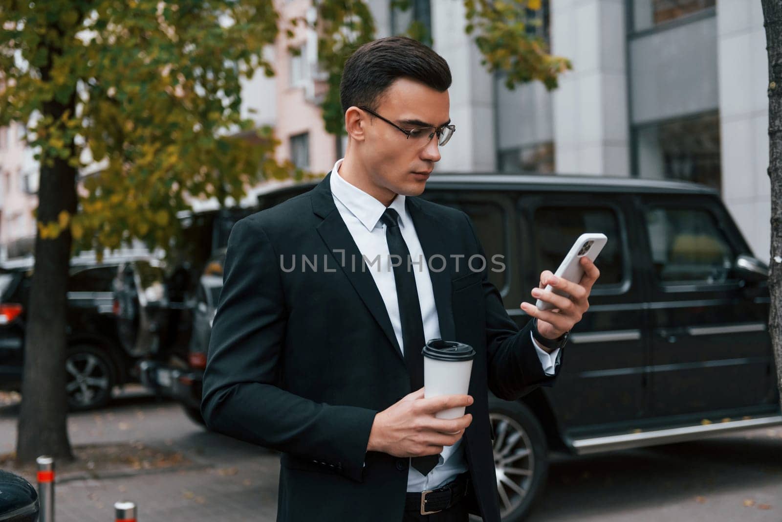 Near black car with phone in hands. Businessman in black suit and tie is outdoors in the city.