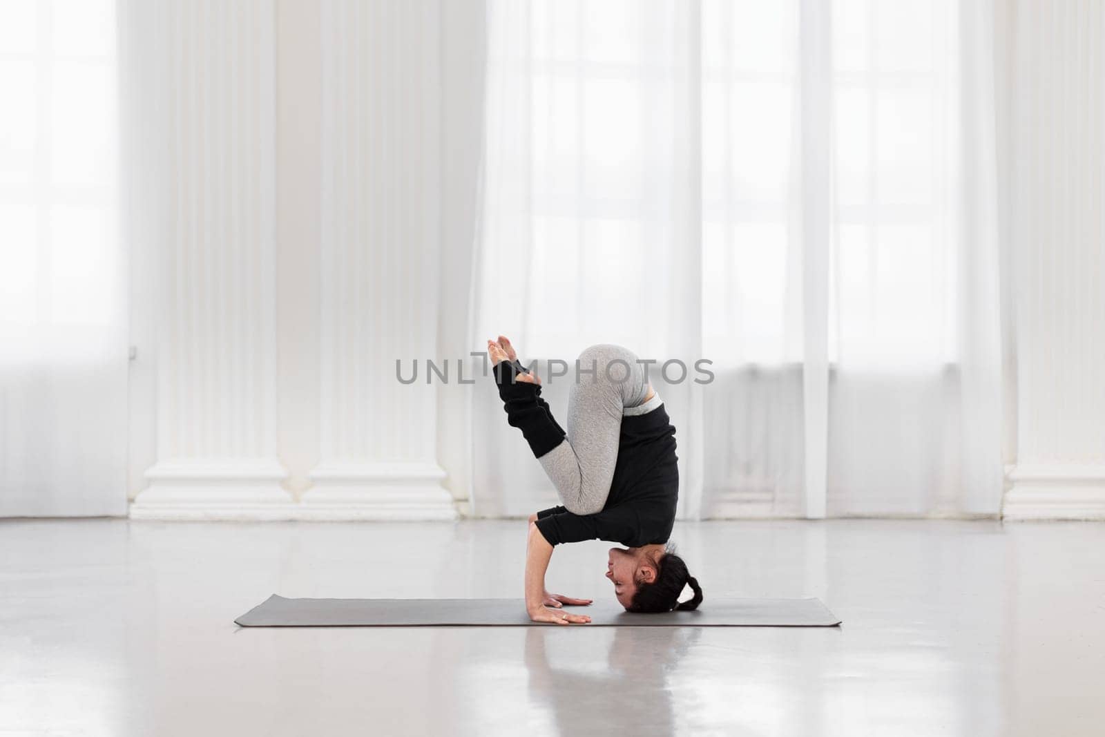 Side view of an athletic woman doing headstand training during yoga meditation in a bright gym. Concept of normalization of all processes in the body, strength, energy and concentration by apavlin