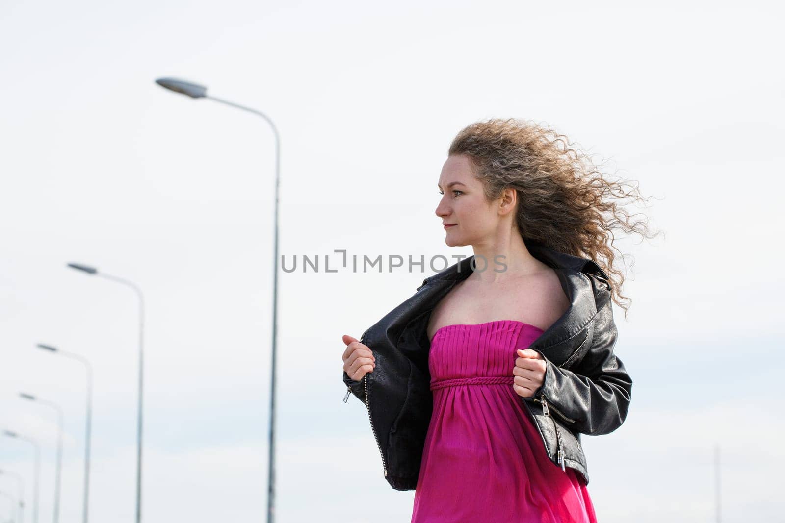 caucasian curly-haired woman in a black leather jacket and a pink dress against the background of a cloudy sky and pillars with lanterns