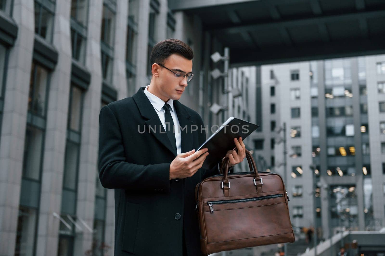 With notepad in hands. Businessman in black suit and tie is outdoors in the city.