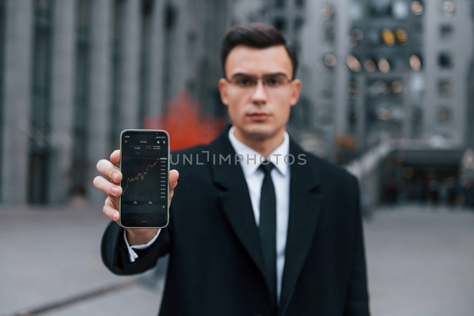 Showing phone with crypto graphs in it. Businessman in black suit and tie is outdoors in the city by Standret