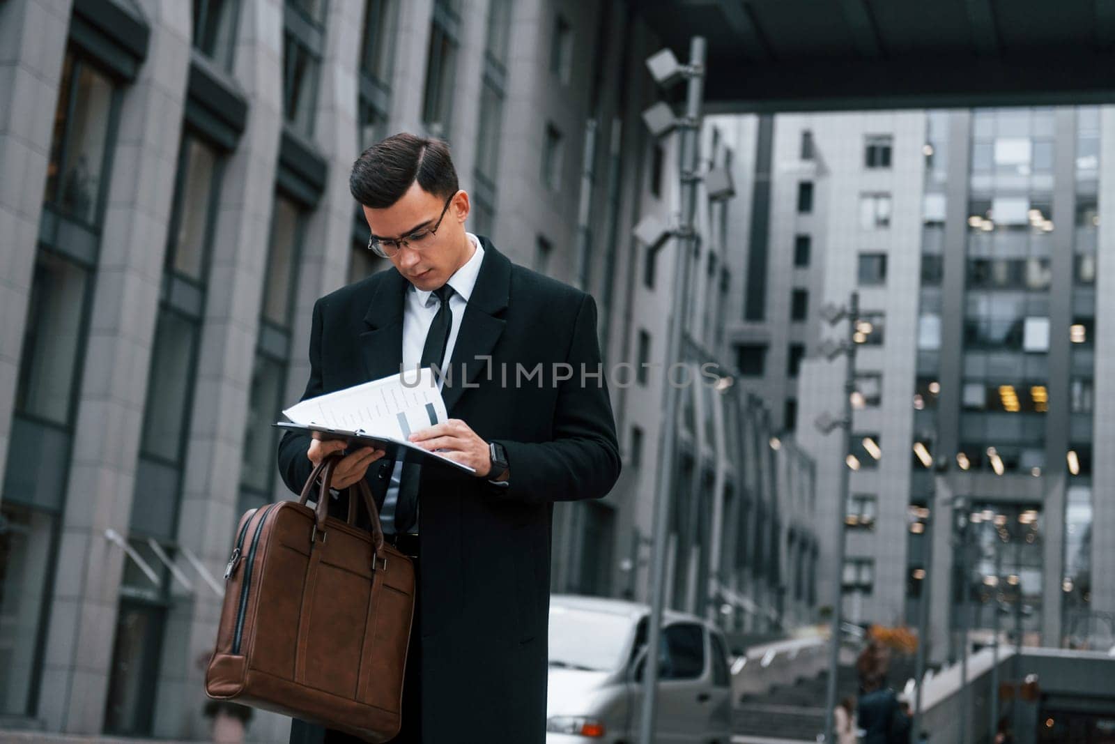 Holding documents. Businessman in black suit and tie is outdoors in the city.