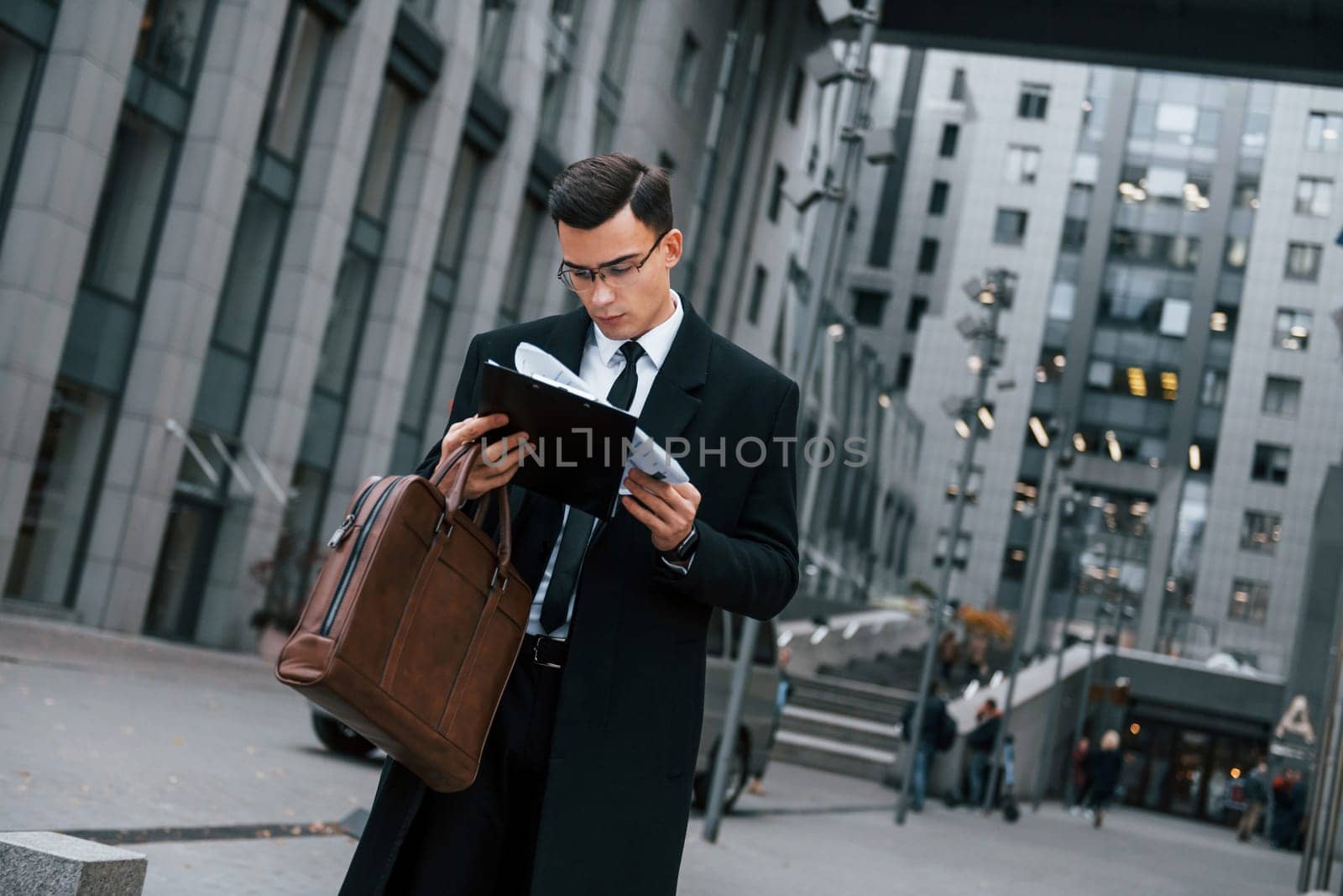 Holding documents. Businessman in black suit and tie is outdoors in the city by Standret