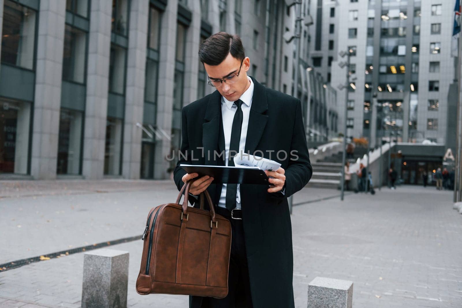 Holding documents. Businessman in black suit and tie is outdoors in the city.