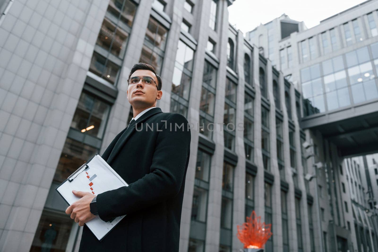 Grey building behind. Businessman in black suit and tie is outdoors in the city.