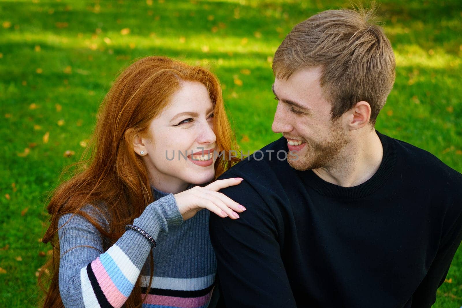 Happy young couple of Caucasian ethnicity man and woman in casual clothes sitting embracing in the park on green grass on a sunny day. Happy relationship between prana and a girl in nature