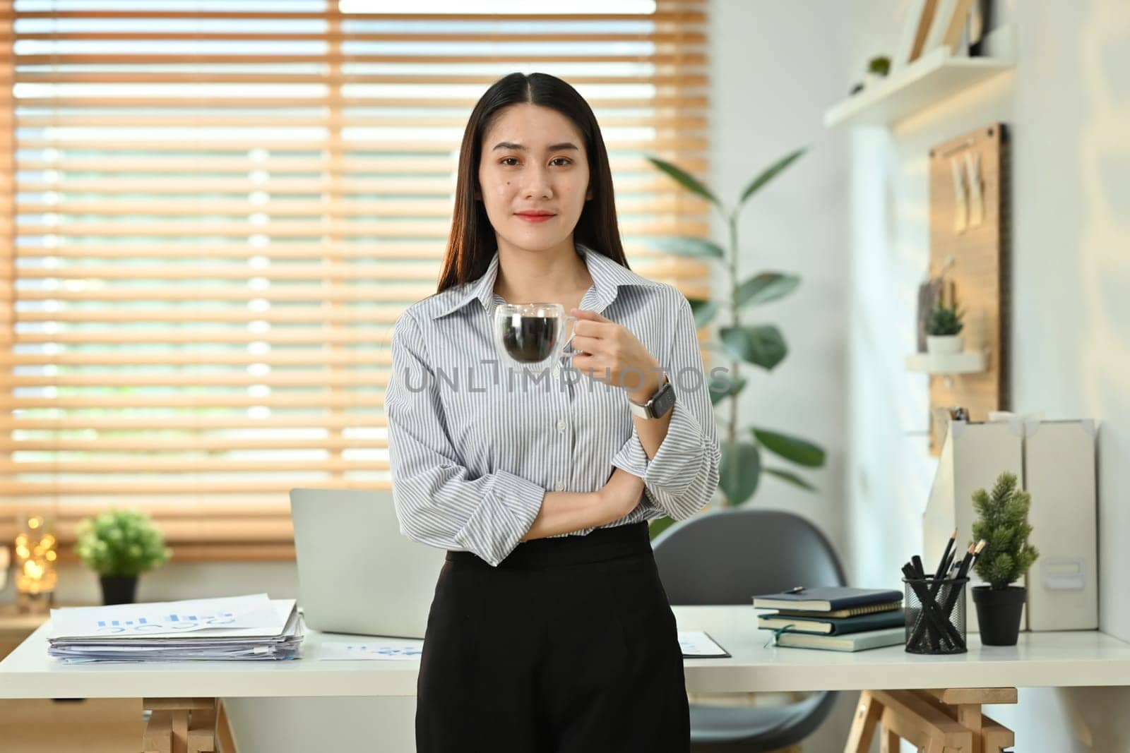 Portrait of millennial female entrepreneur holding cup of coffee standing in front of her desk and smiling at camera.