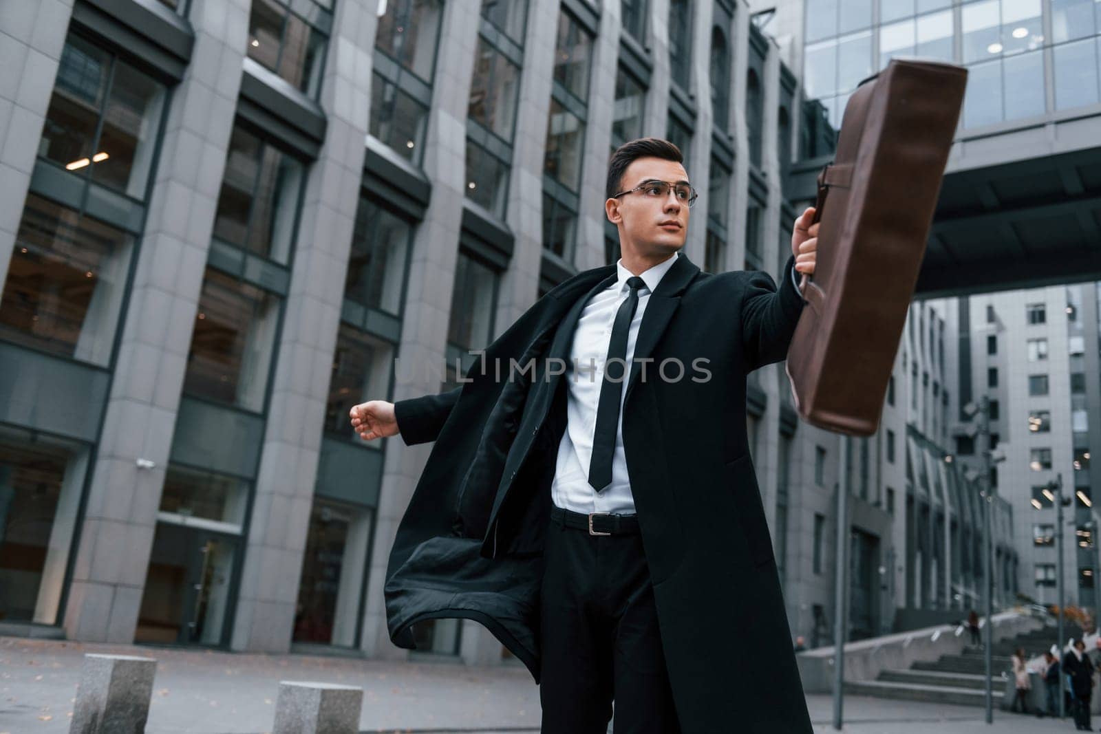 Grey building behind. Businessman in black suit and tie is outdoors in the city.