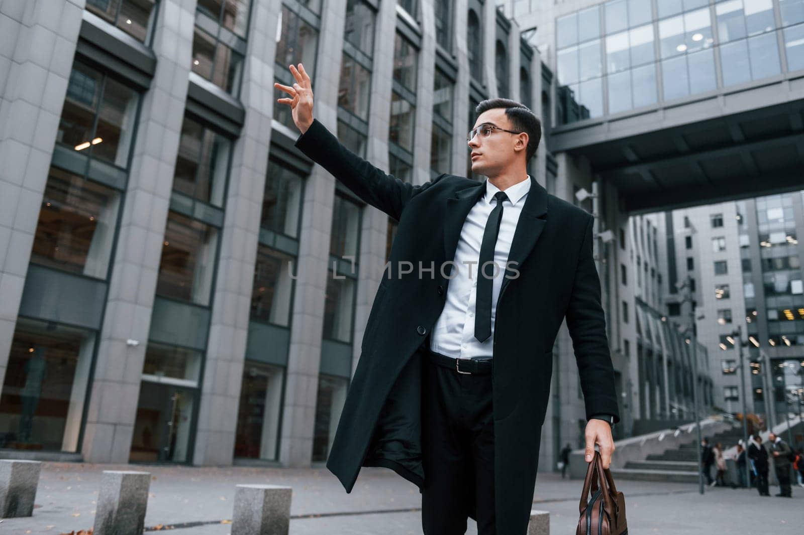 Raises his right hand. Businessman in black suit and tie is outdoors in the city.