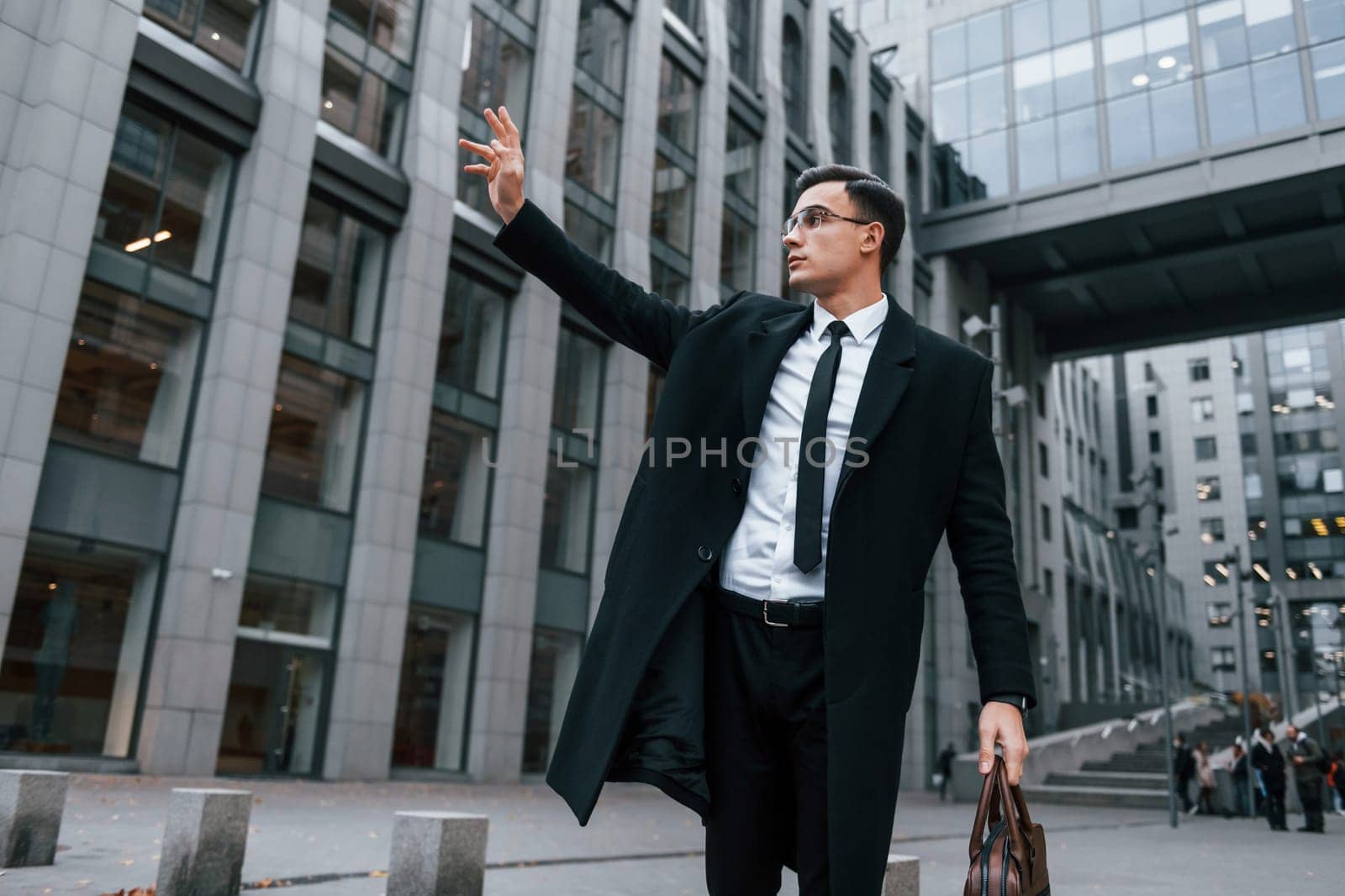 Raises his right hand. Businessman in black suit and tie is outdoors in the city by Standret