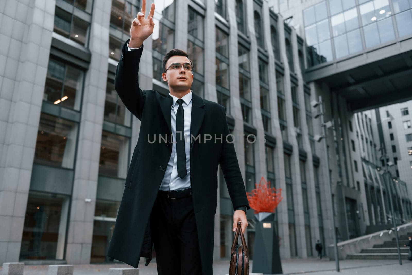 Raises his right hand. Businessman in black suit and tie is outdoors in the city.