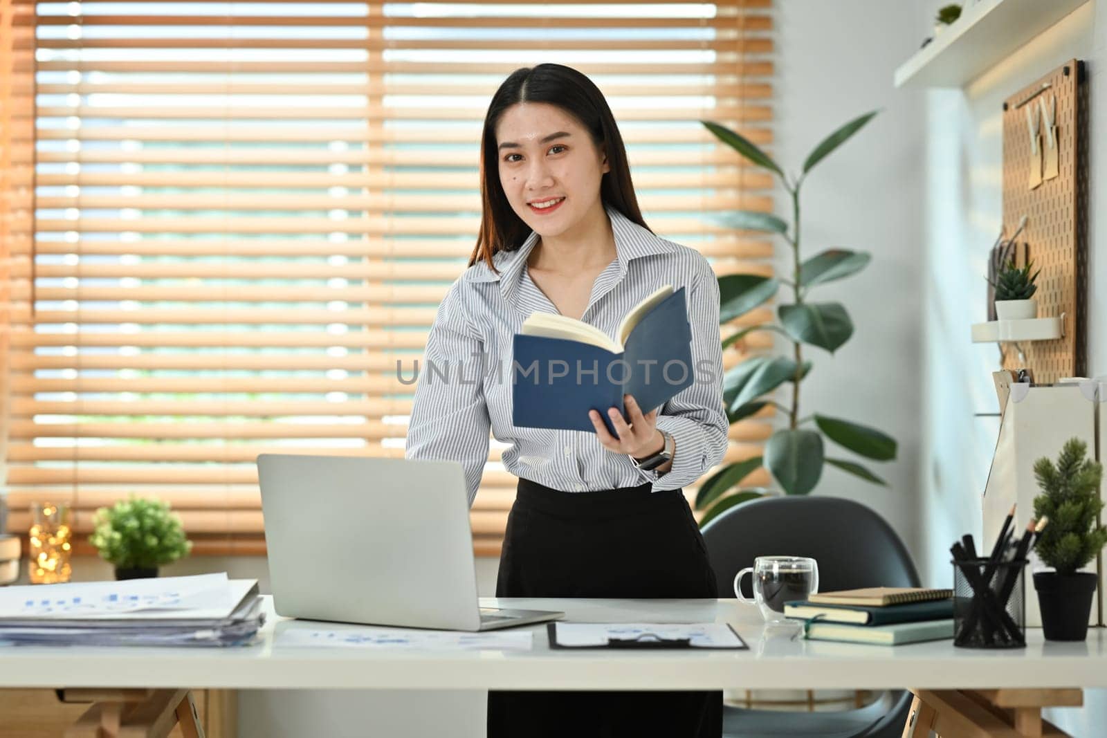 Beautiful young female entrepreneur holding diary planner standing at her working desk and using laptop computer by prathanchorruangsak