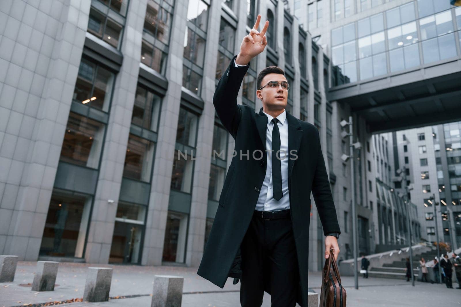 Raises his right hand. Businessman in black suit and tie is outdoors in the city by Standret