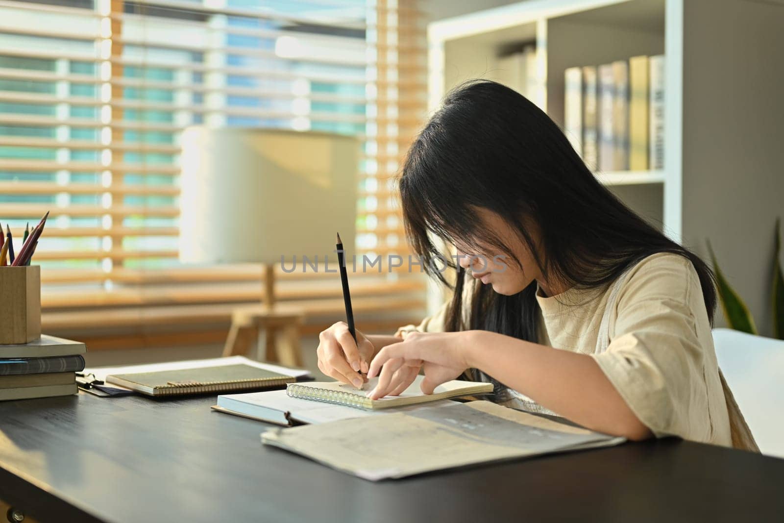Focused teenaged girl doing homework, writing in notebook while sitting at table in living room.