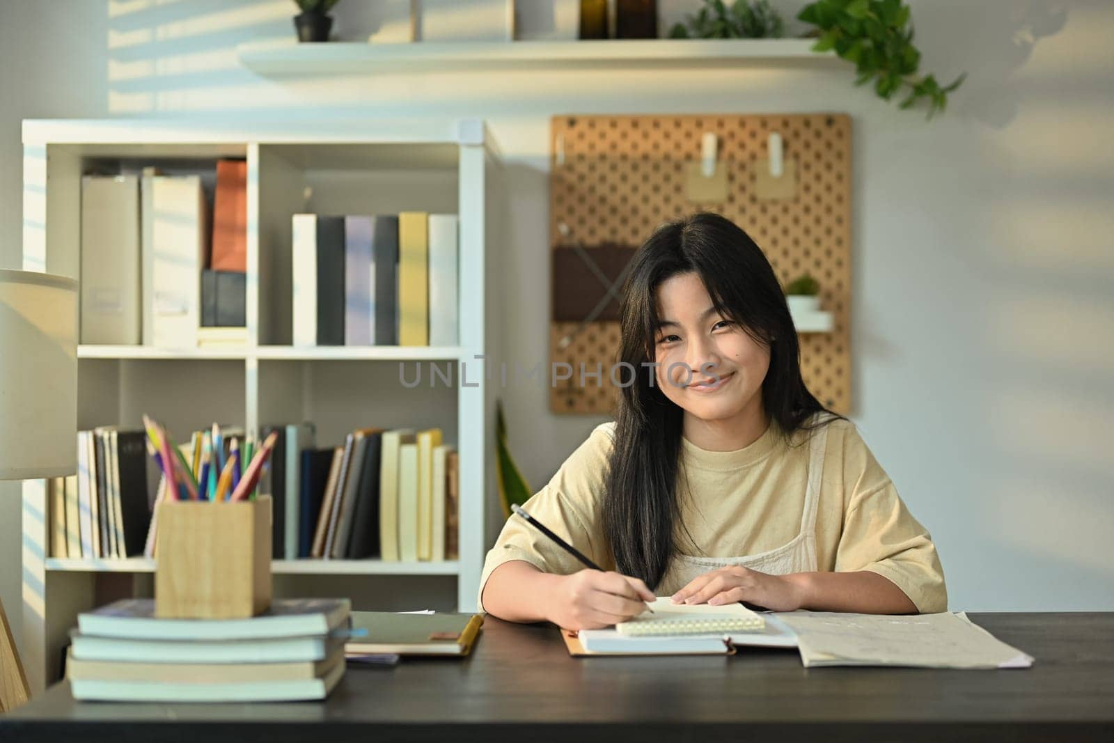 Cute teenager girl doing homework or preparing for exam at desk in living room. Homeschooling and education concept.