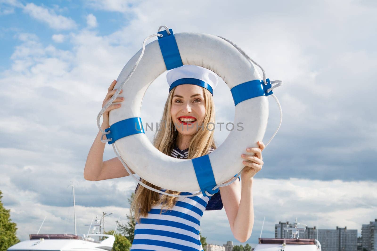 Young happy woman of Caucasian appearance in a blue striped dress standing on a yacht posing with a lifebuoy in her hand, against the background of a blue sky with clouds on a summer sunny day