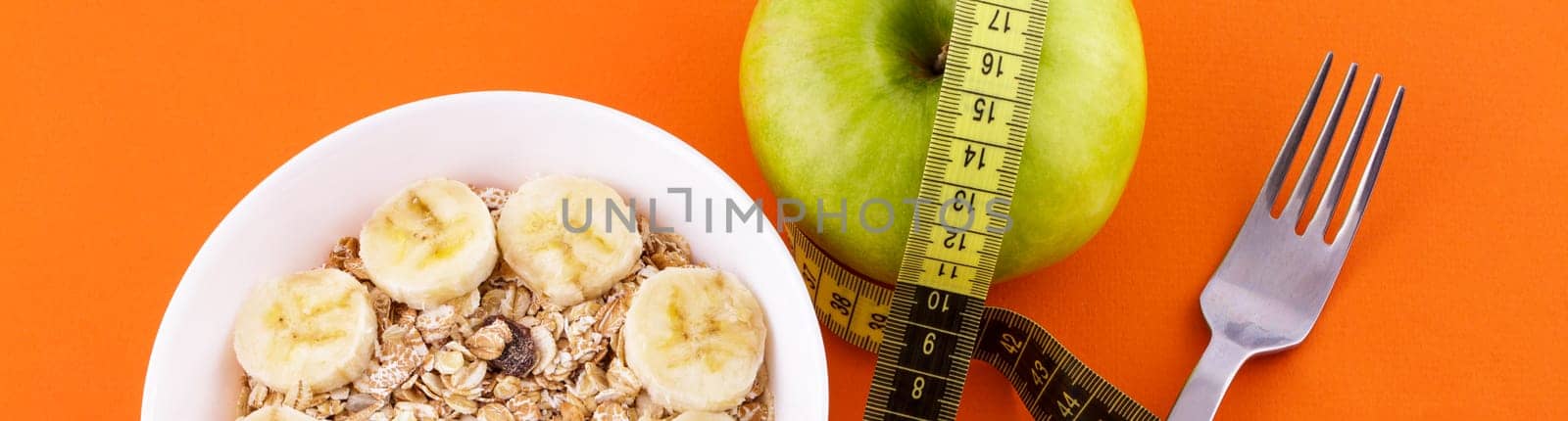 muesli with banana in a white plate on an orange background, a fork and a green apple with a yellow measuring tape. Healthy food and diet concept