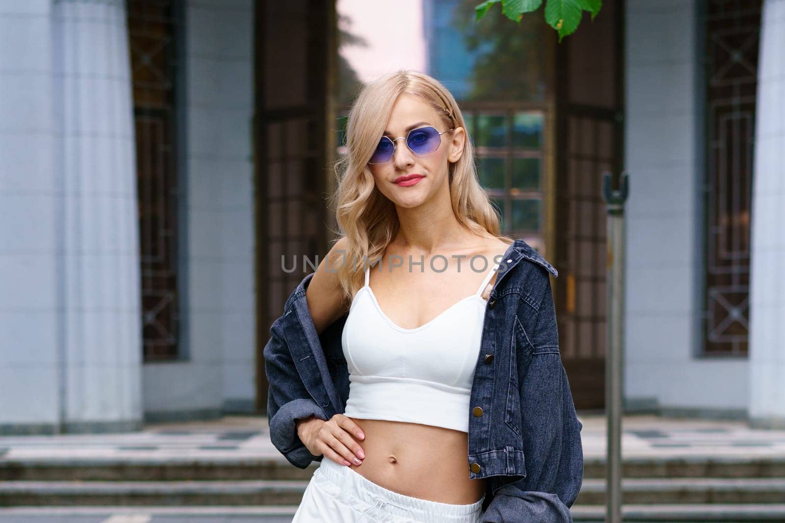 Young beautiful woman of Kakaz nationality poses in the afternoon on the street of the city in a white tracksuit and blue jeans. Confident blonde woman in sunglasses in summer