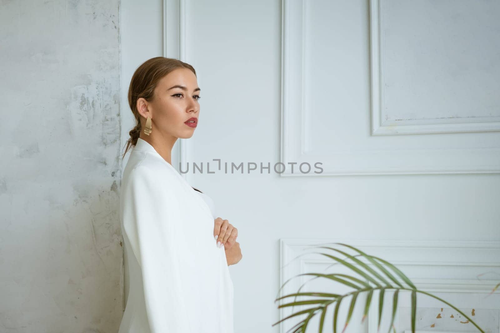 portrait of a beautiful young blonde woman with large gold earrings and a white jacket on the background of the white interior of the living room. The model is taken in daylight and looks away.