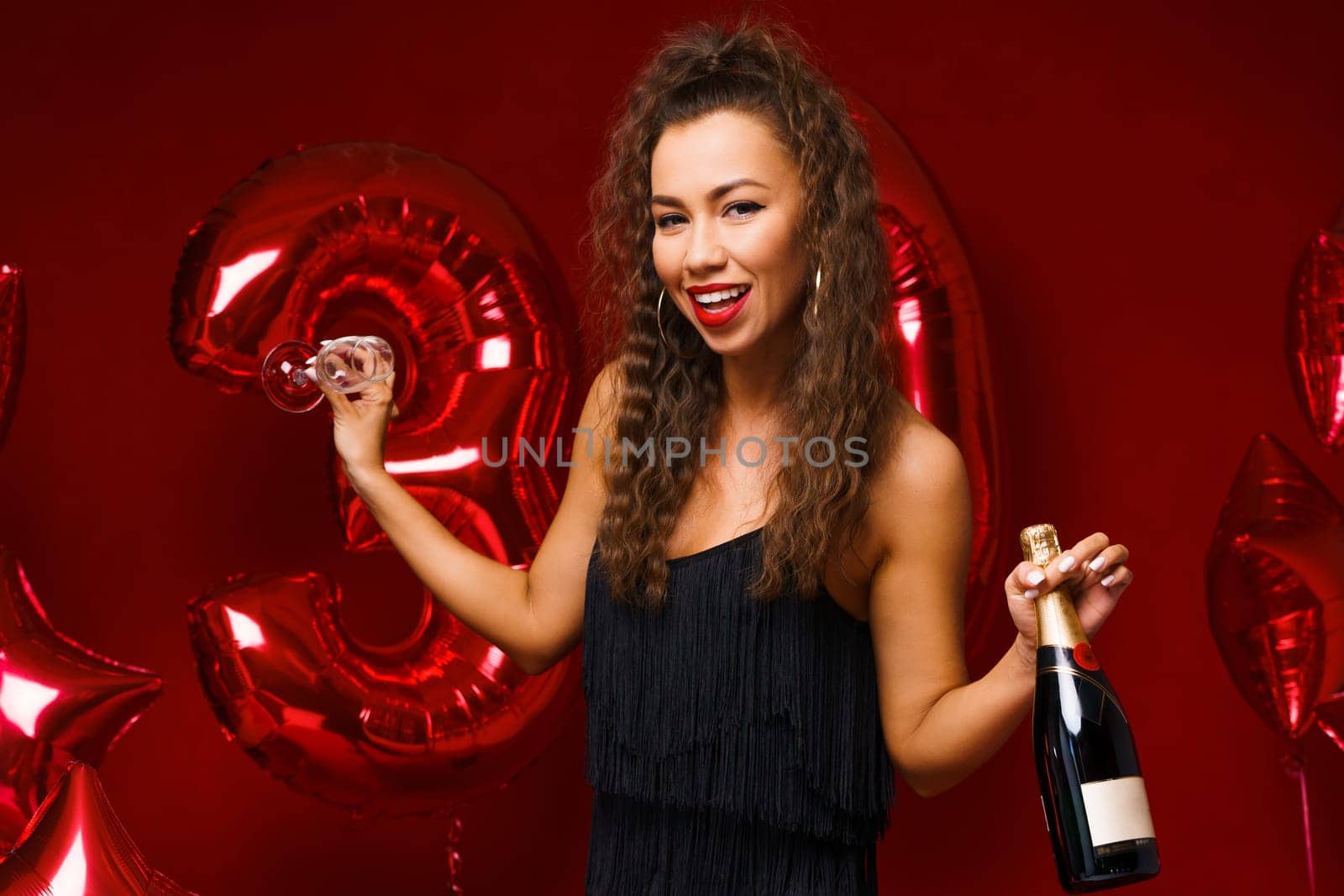 Beautiful woman posing on a red background with balloons holding a bottle of champagne in her hand. Cheerful girl of Caucasian appearance with wavy hair and bright makeup celebrates her 30th birthday