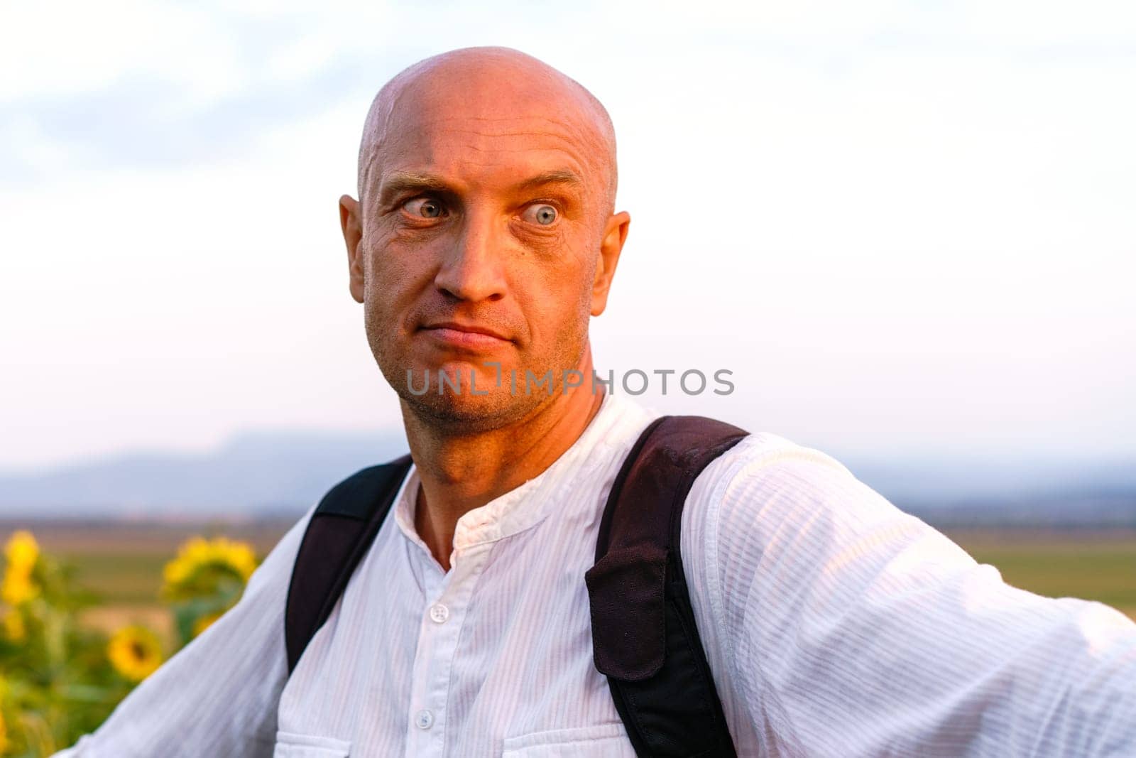 Close-up of a cheerful young man in a white t-shirt with a backpack stands and smiles in a field of sunflowers. Cute caucasian bald man outdoors during sunset
