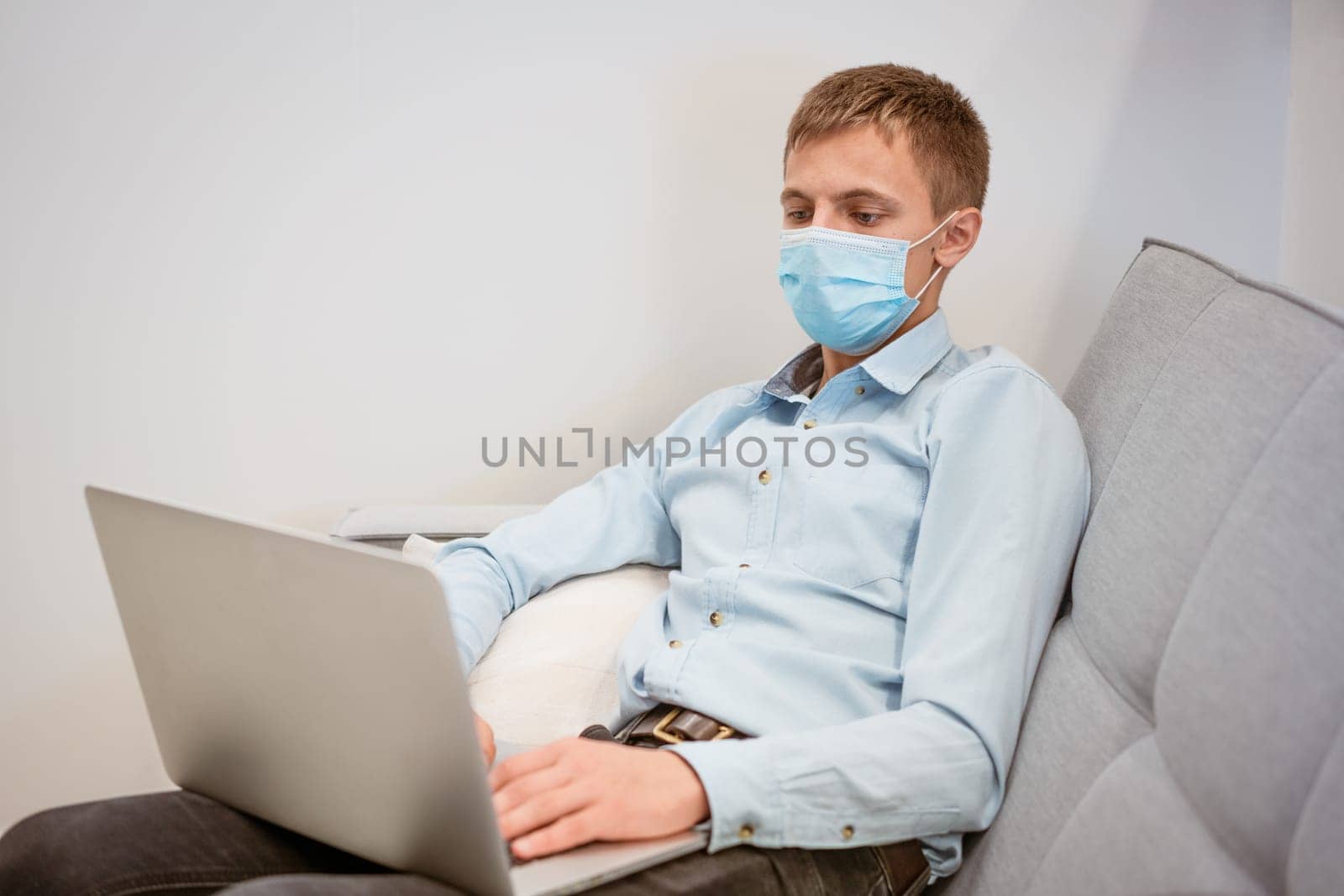 a young guy of European appearance in a light shirt, sits in a protective medical mask and works on the couch via video communication, remote work during a global pandemic