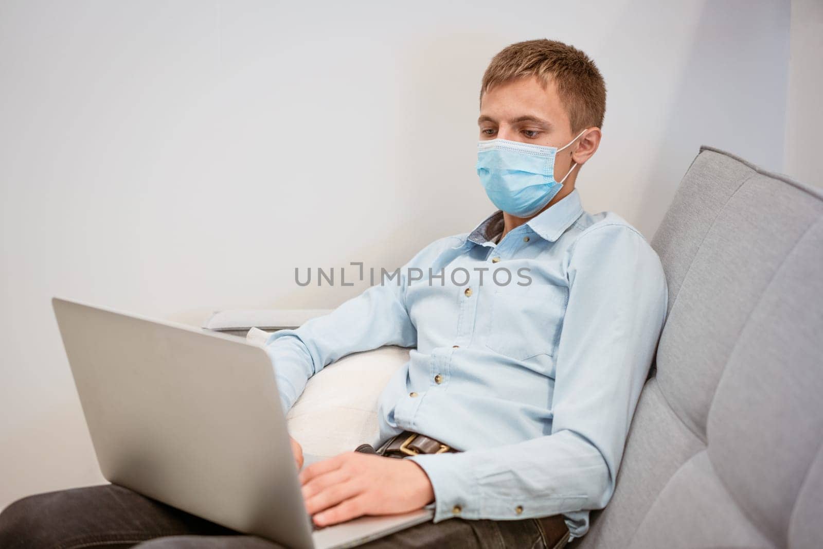 a guy sitting in a protective mask works on the couch via video communication by EkaterinaPereslavtseva